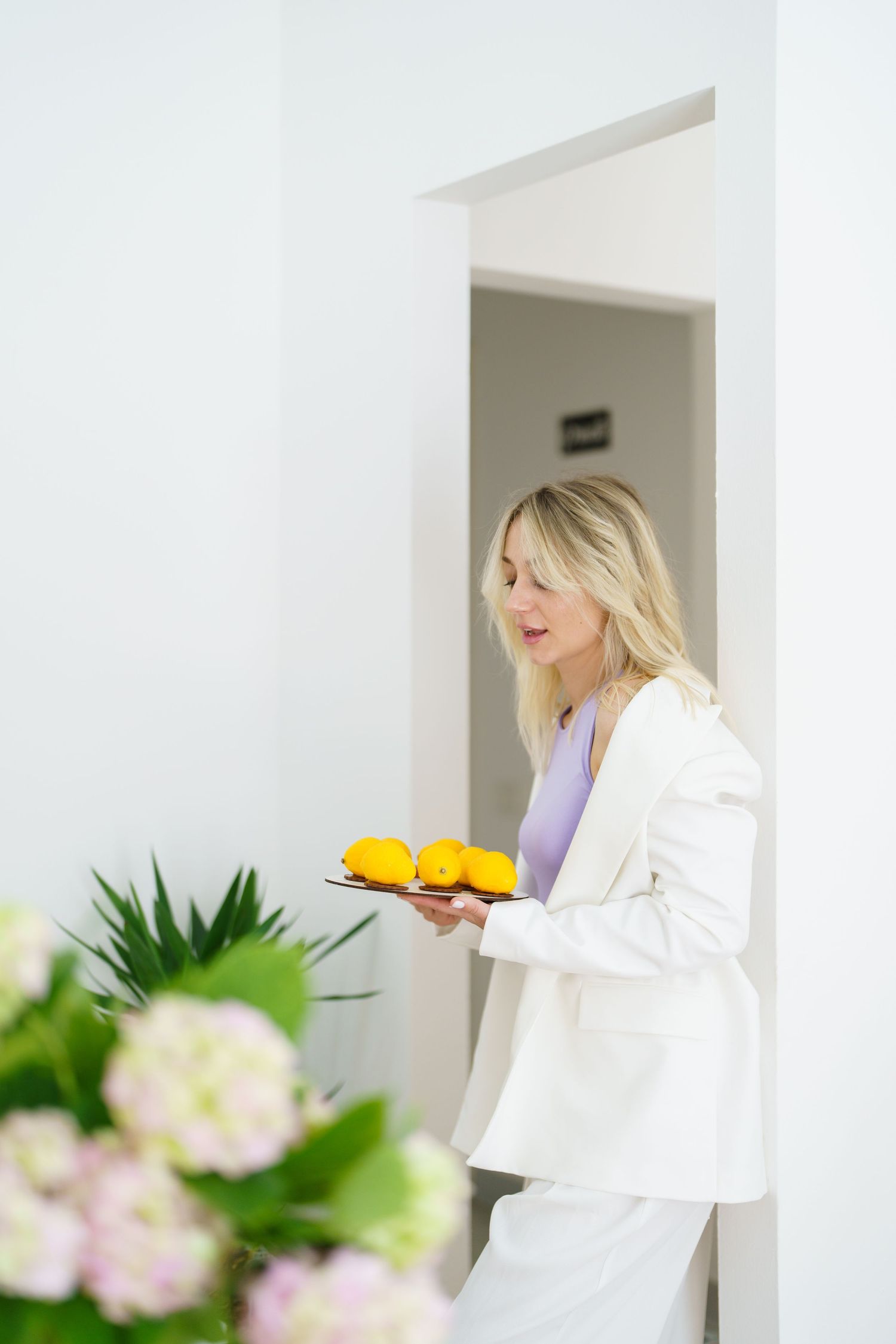 Elegant blonde woman in white suit holding a tray of yellow lemons in a bright modern interior.