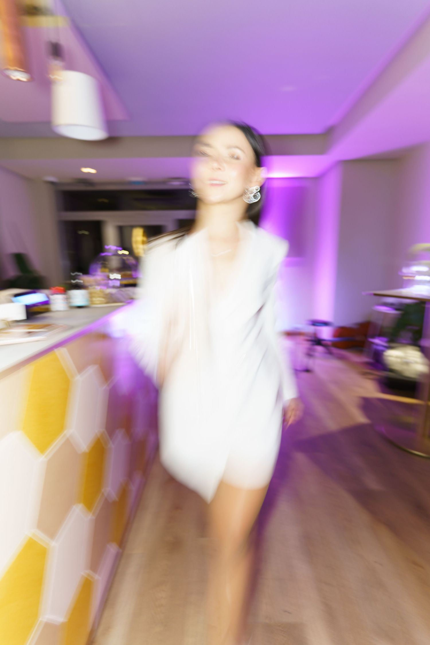 A woman in a white mini dress and fur stole poses elegantly in a purple-lit modern bar setting.