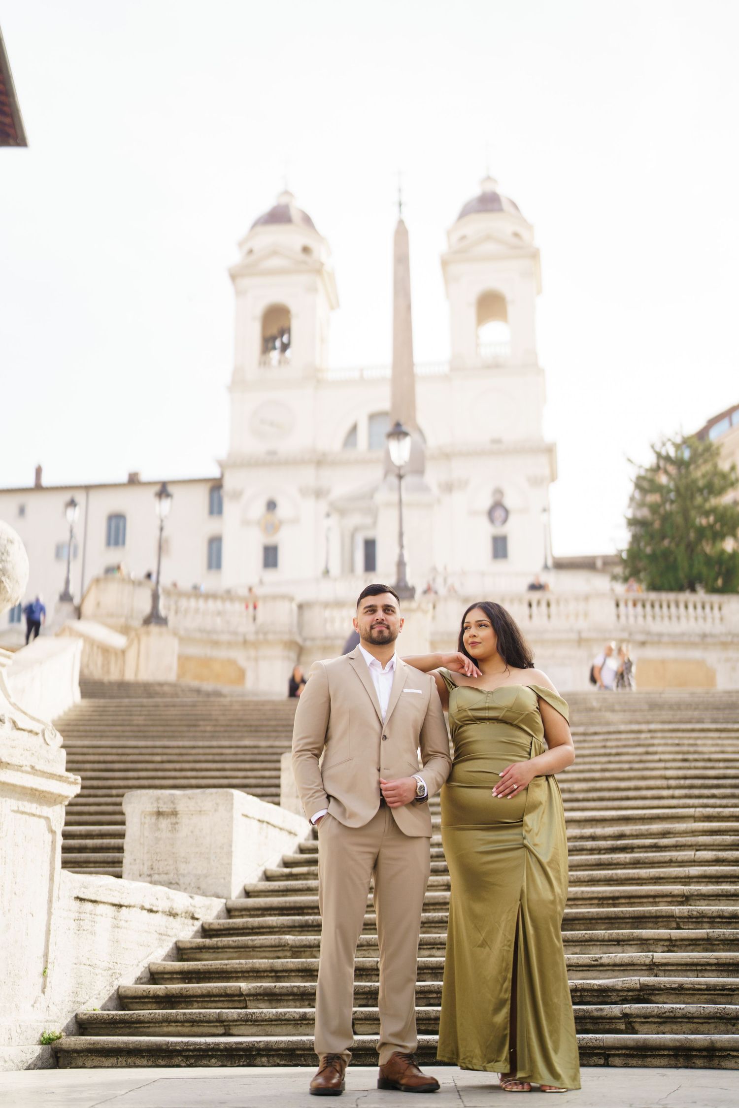 Elegant couple posing on the Spanish Steps in Rome, woman in green gown, man in tan suit, Trinità dei Monti church behind.