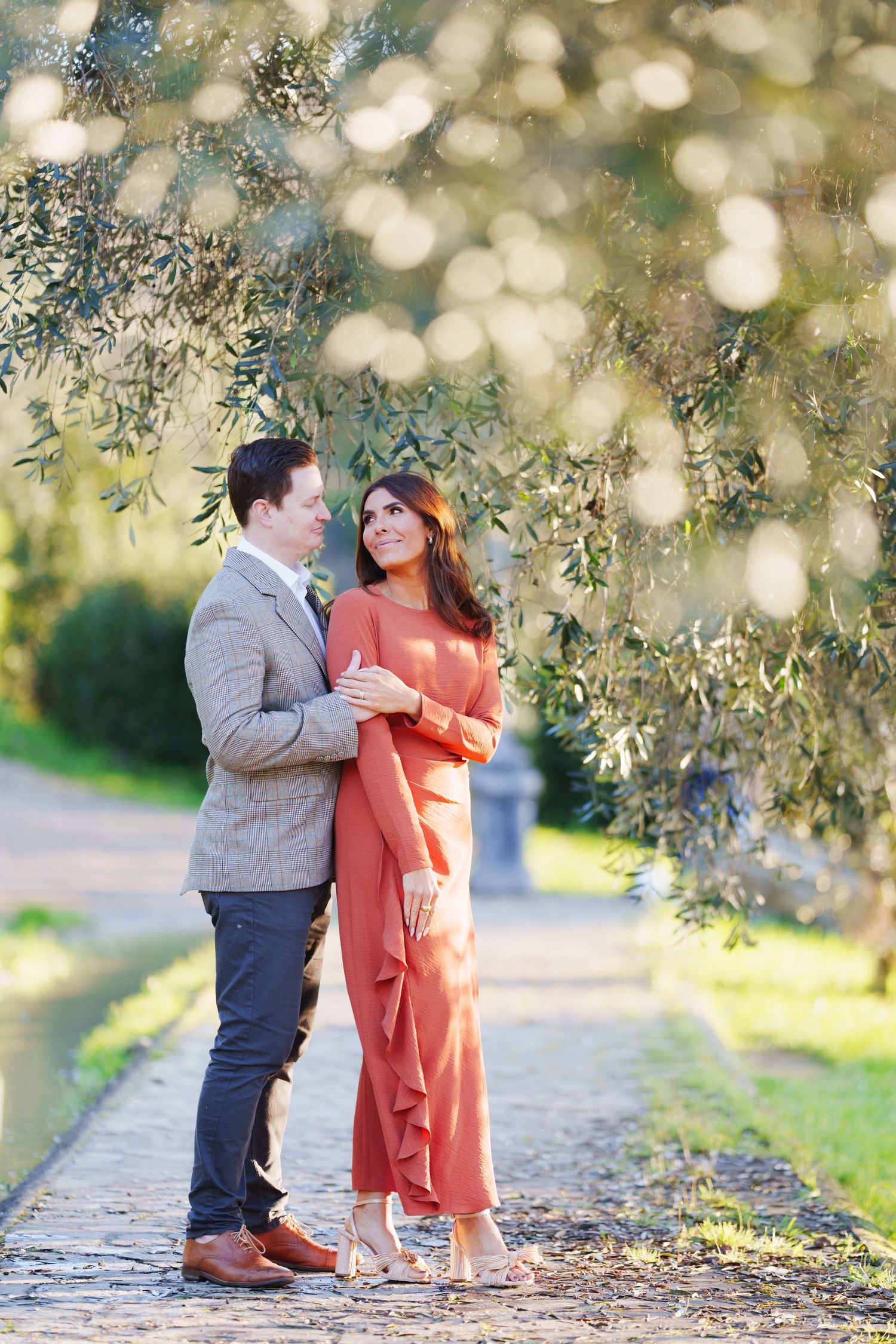 Couple in elegant attire embracing under olive tree branches in a sunlit garden pathway.