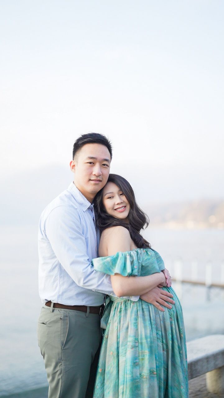 A couple embraces by a lakeside, the woman in a teal maternity dress and man in a light blue shirt, smiling warmly.