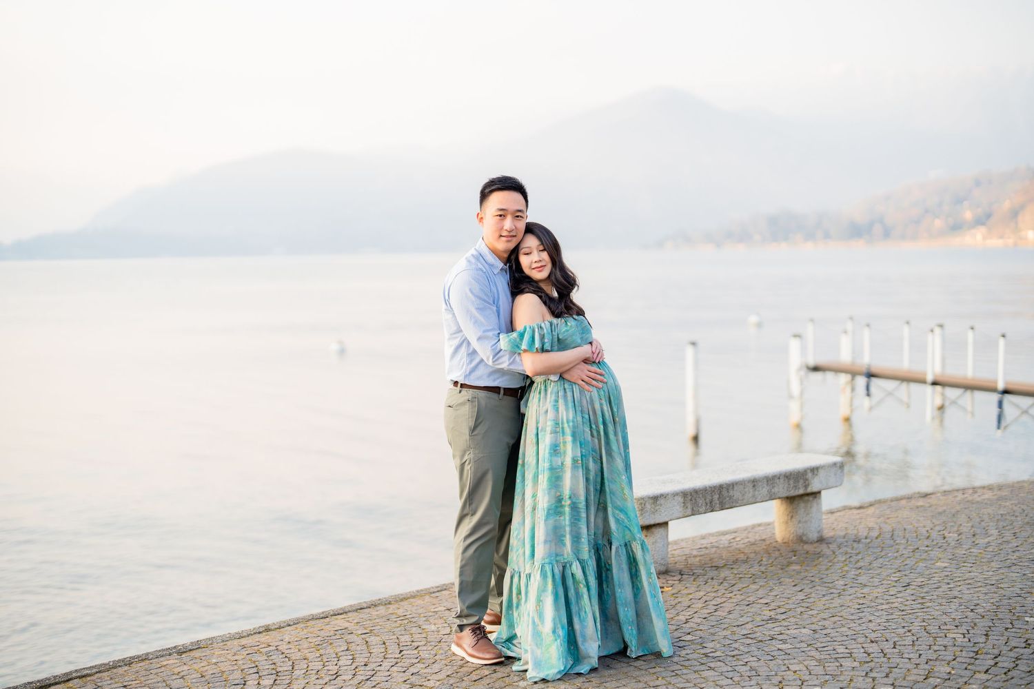 Couple embracing by a lakeside promenade with mountain backdrop during a maternity photoshoot at golden hour.