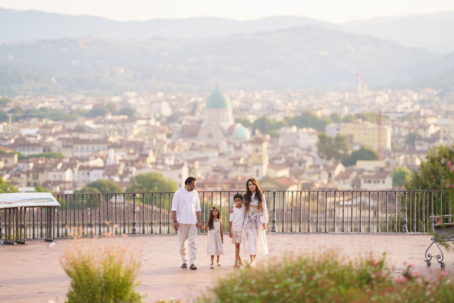 Family of four in white outfits strolling at a scenic overlook with Florence cityscape and dome in the background.