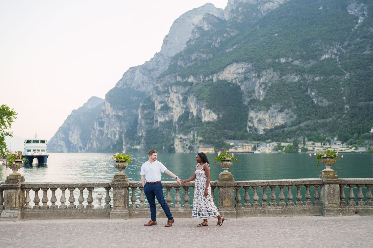 Couple holding hands and walking on elegant Lake Garda terrace with mountains and water views.