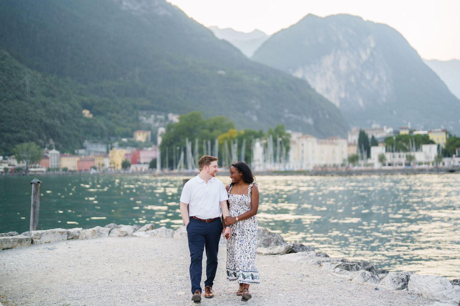 Happy interracial couple walking together by a sparkling lake with mountains and town behind them.