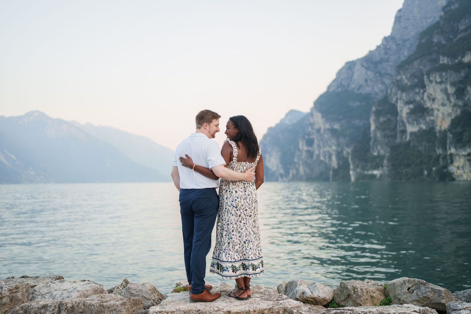 Couple standing close together on rocks by a tranquil mountain lake with misty cliff backdrop.