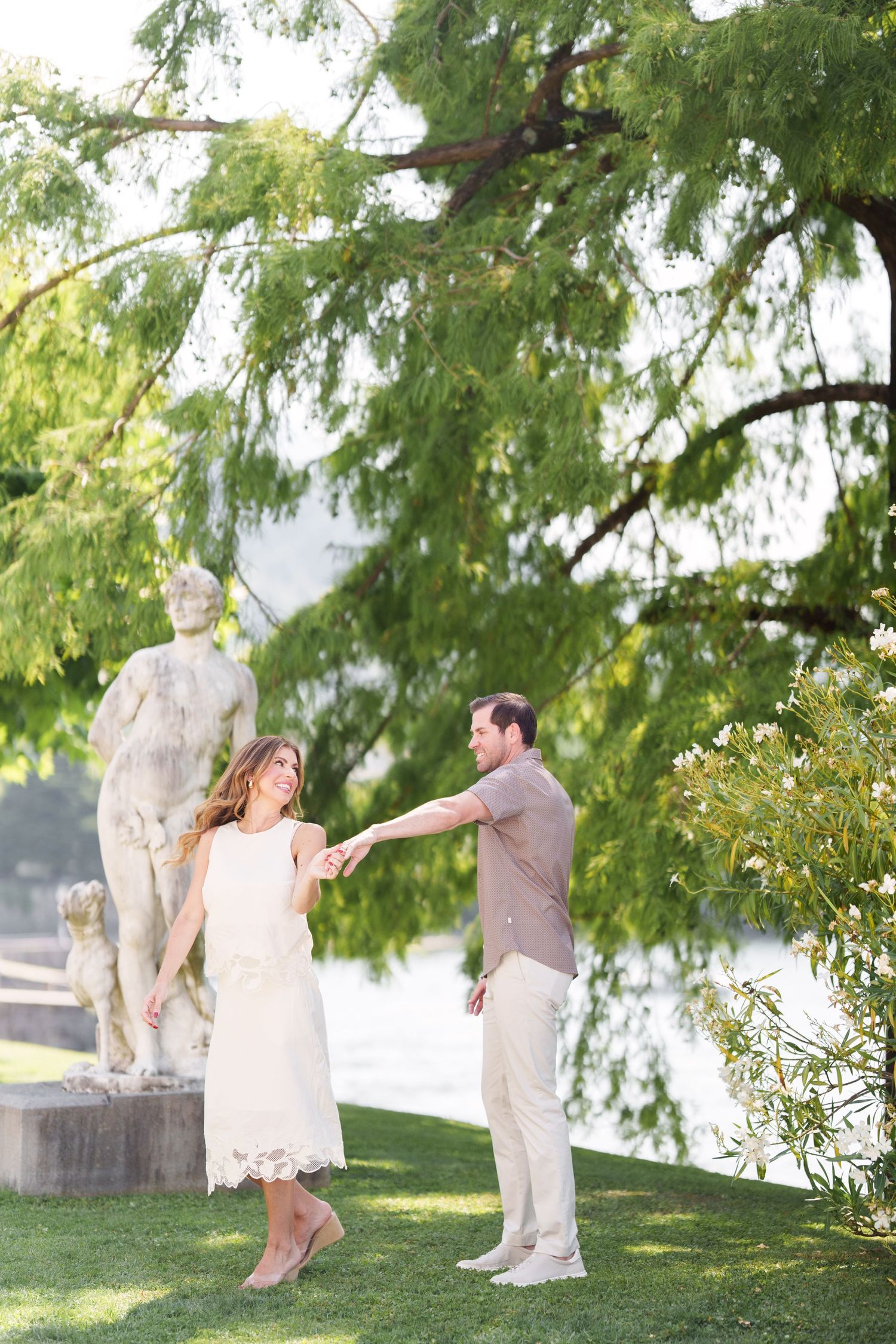 Couple dancing on green lawn near classical statues and a serene waterway, surrounded by lush trees in golden light.