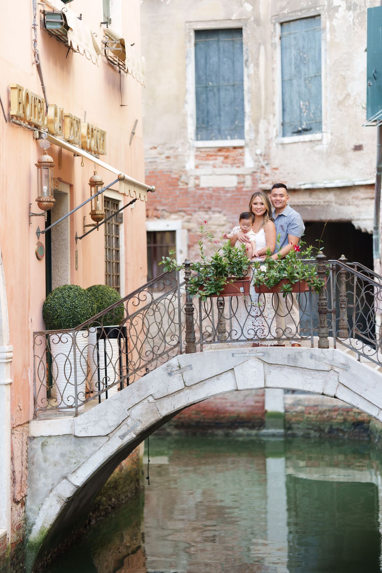 Family posing on a charming stone bridge over a Venice canal, surrounded by colorful flowers and historic buildings.