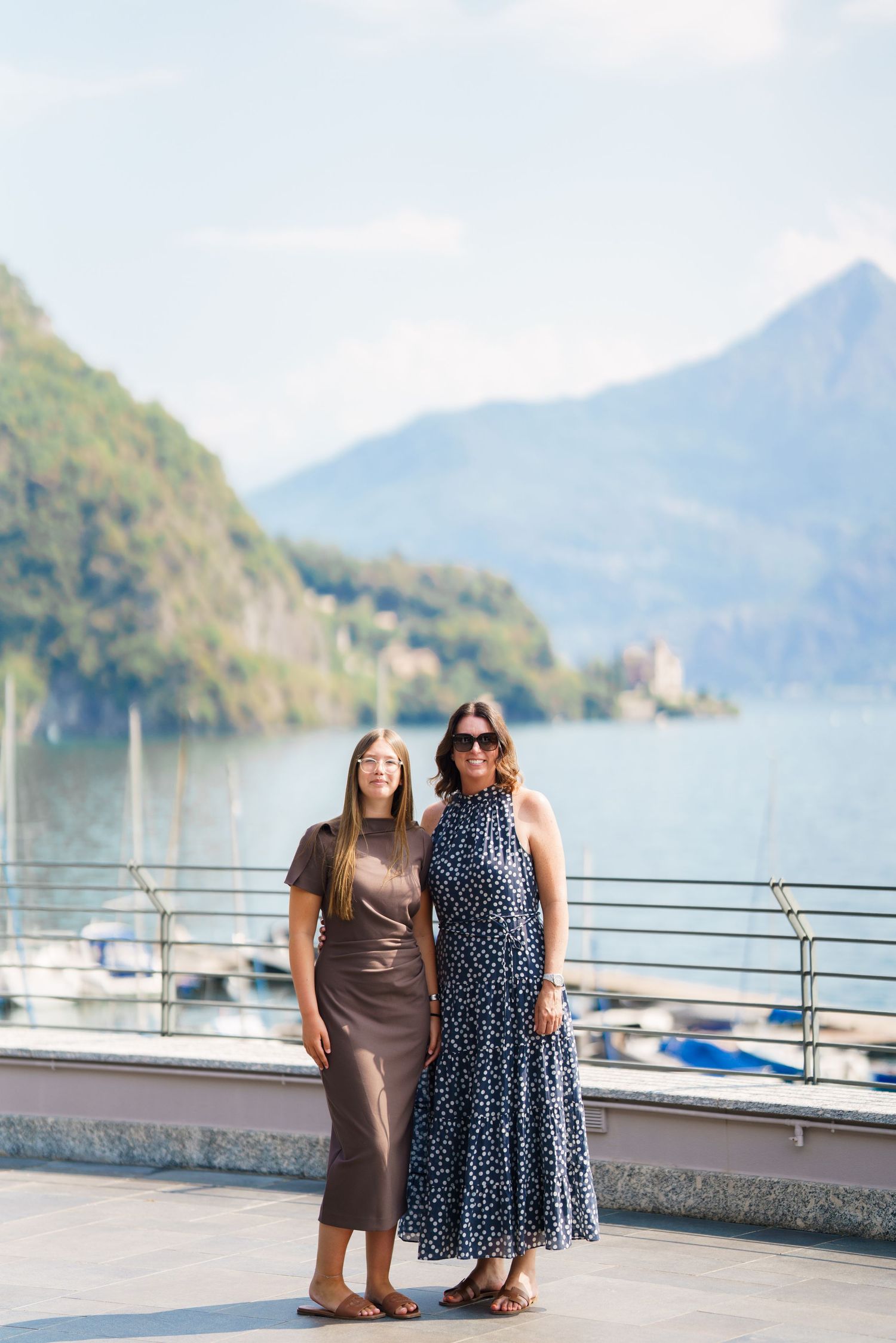 Two women posing on a lakeside terrace with mountain views, sailboats, and blue skies in the background.