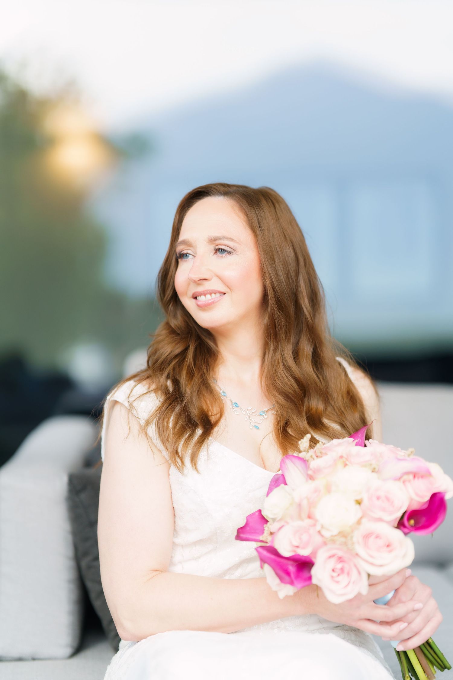 Smiling bride in white dress holding pink and white bouquet of roses while seated outdoors.