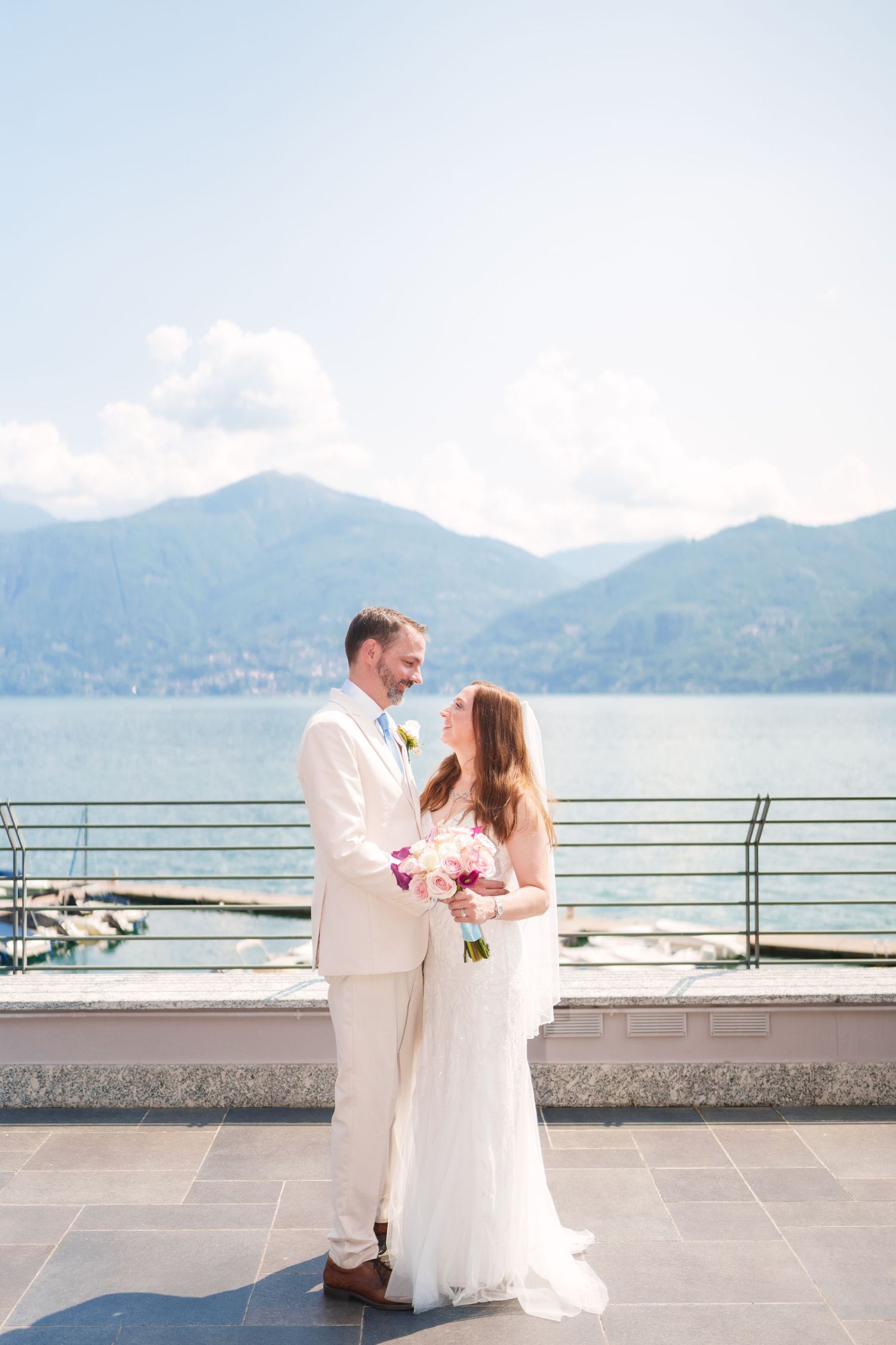 Bride and groom share a romantic moment on a lakeside terrace with mountain views, bride holding a pink bouquet.