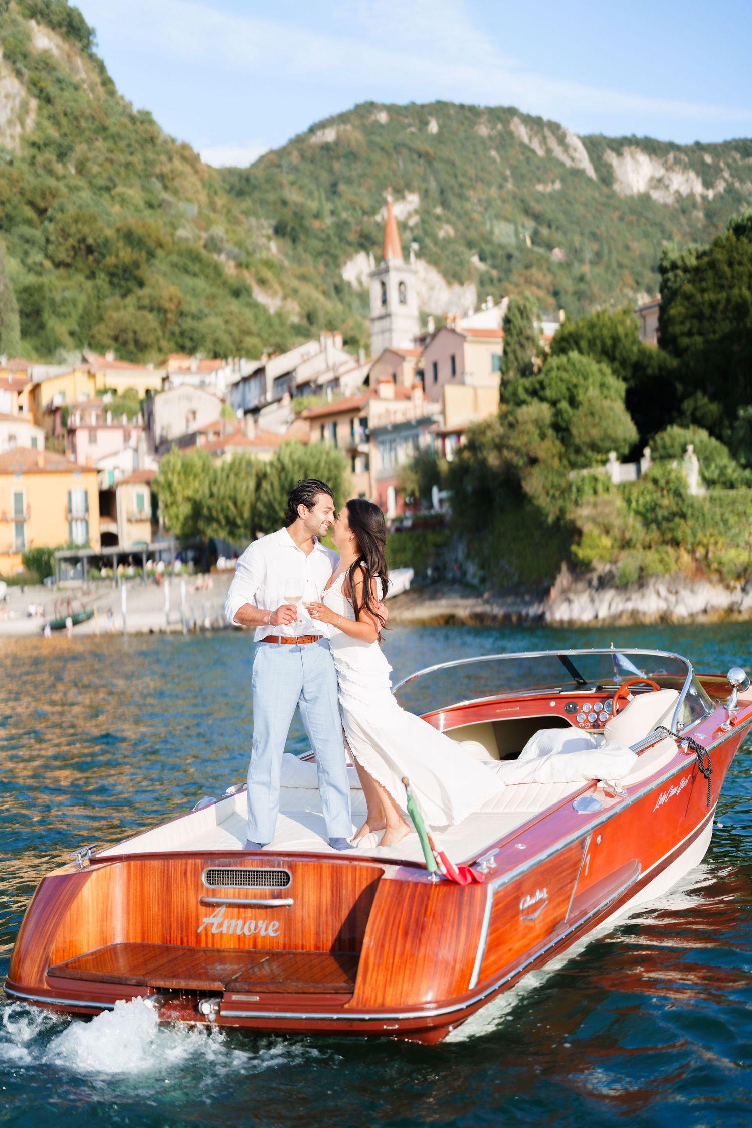 Couple sharing a romantic kiss on a classic wooden Riva boat on Lake Como, Italy, with a picturesque village backdrop.