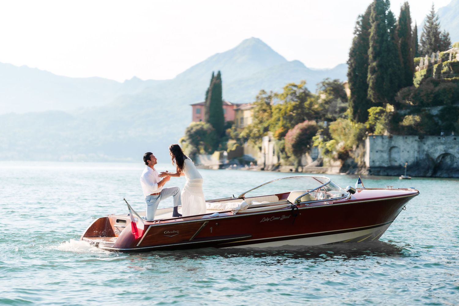 Couple in white attire on a classic wooden speedboat on a serene lake with mountains and villa backdrop.