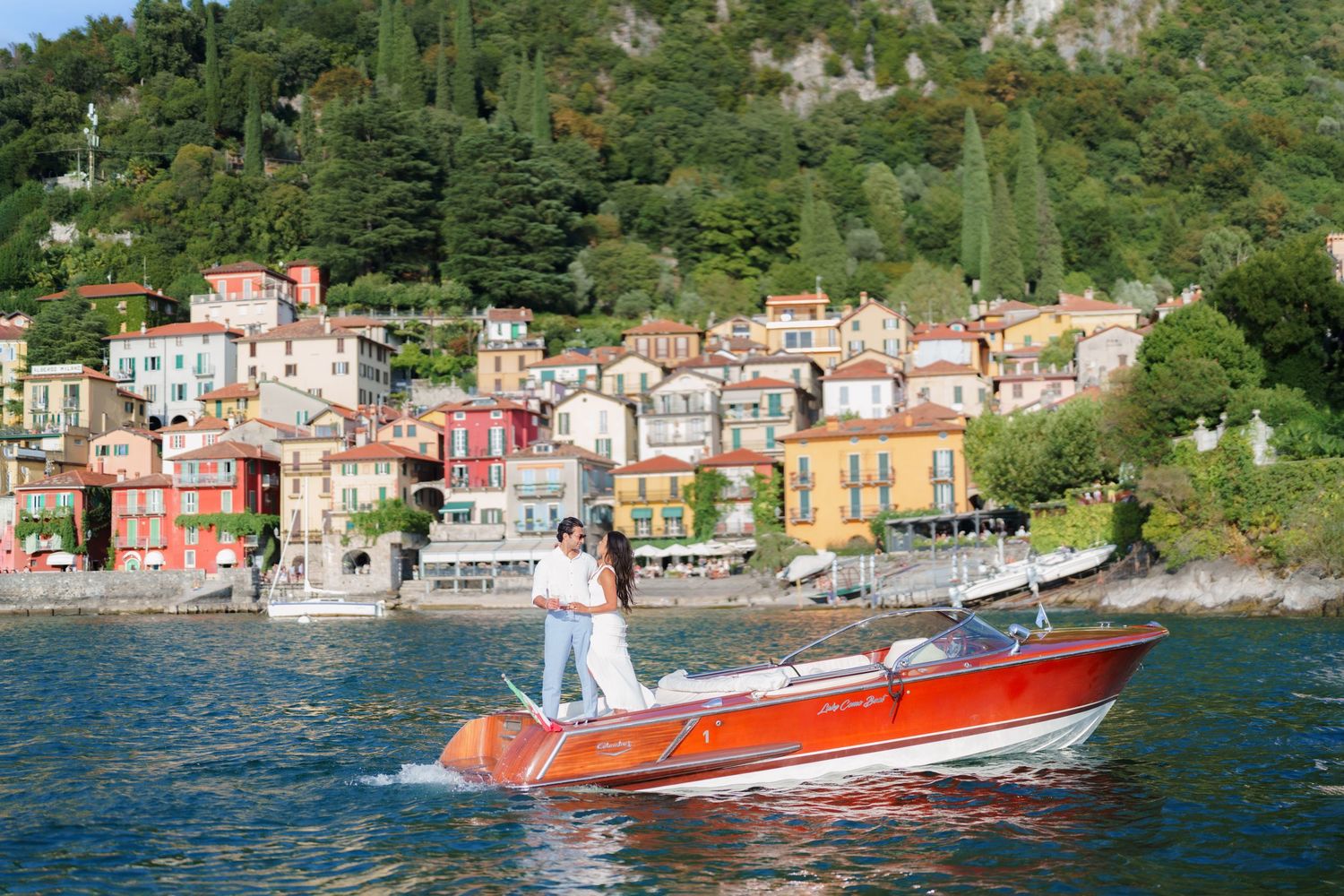 A couple stands on a red wooden speedboat on Lake Como with colorful Italian village buildings and lush green hills behind them.