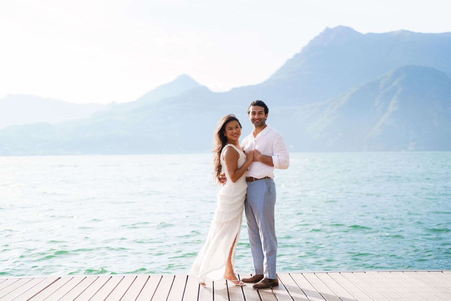 Couple posing on a wooden dock by a turquoise lake with mountain backdrop, woman in white dress, man in white shirt.