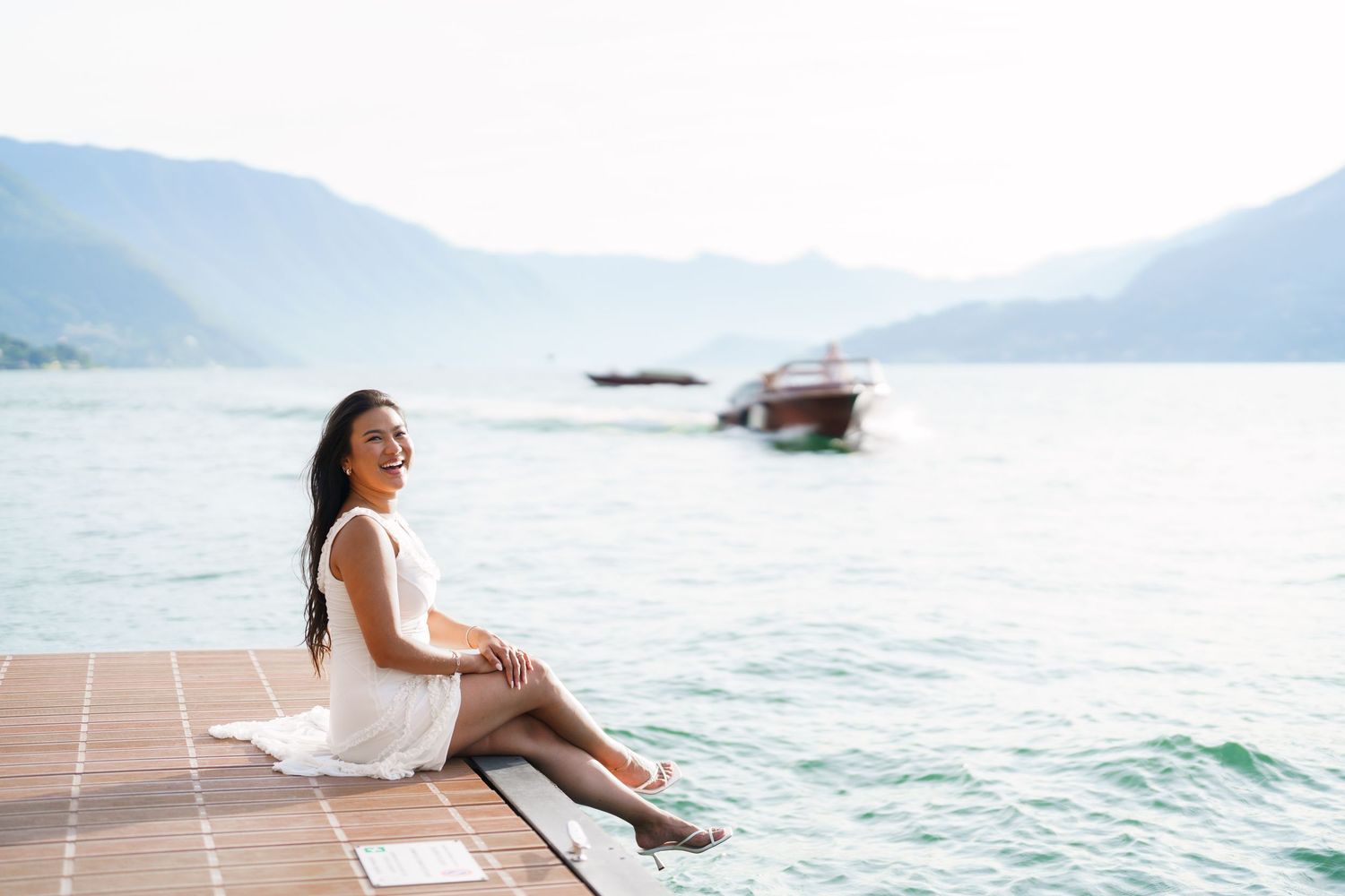 Woman in white dress sitting on wooden dock by turquoise lake with mountains and boats in background.