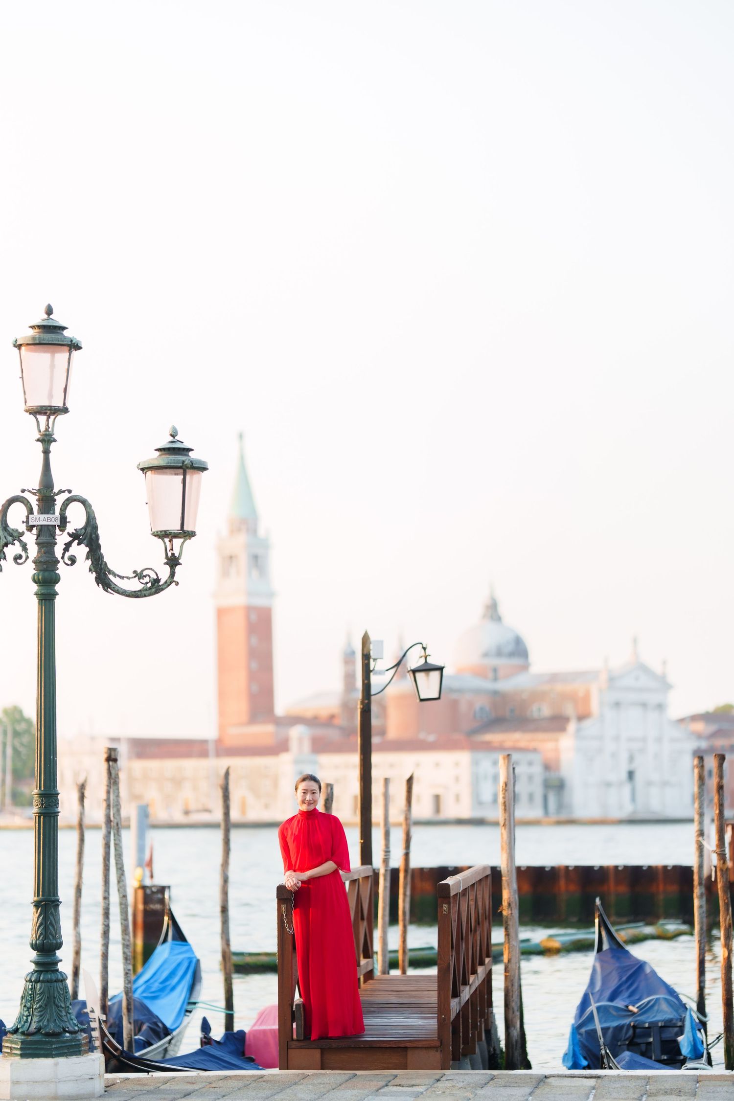 Woman in red dress standing on Venice waterfront with gondolas and San Giorgio Maggiore church in background.