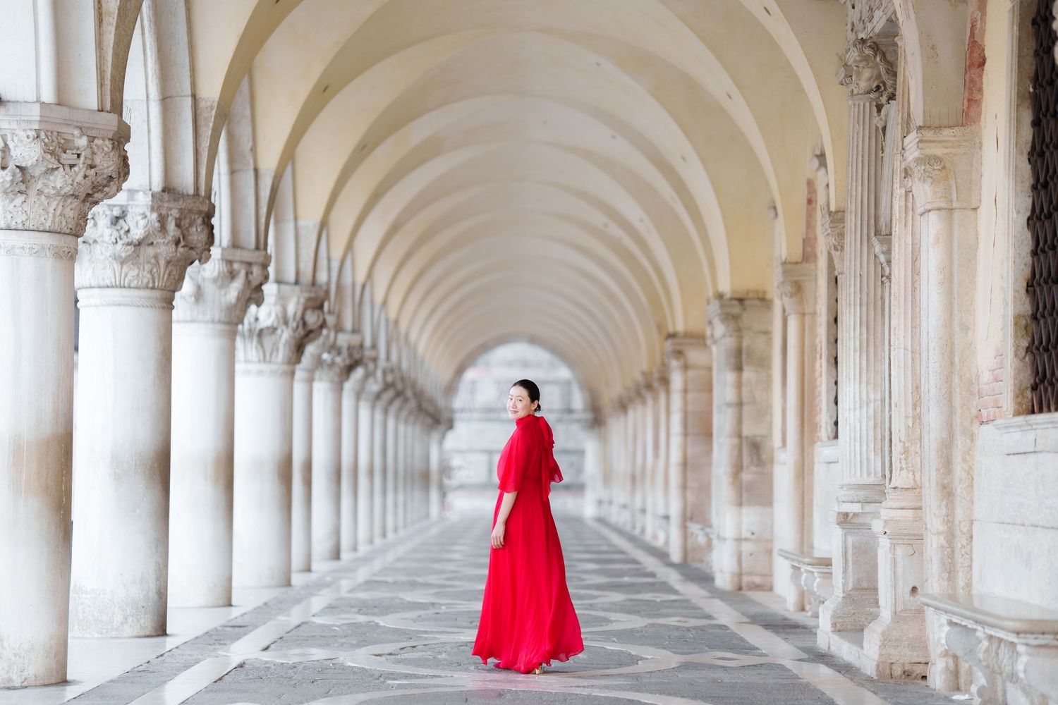 Woman in red gown stands in elegant white marble colonnade arcade in Venice, Italy.
