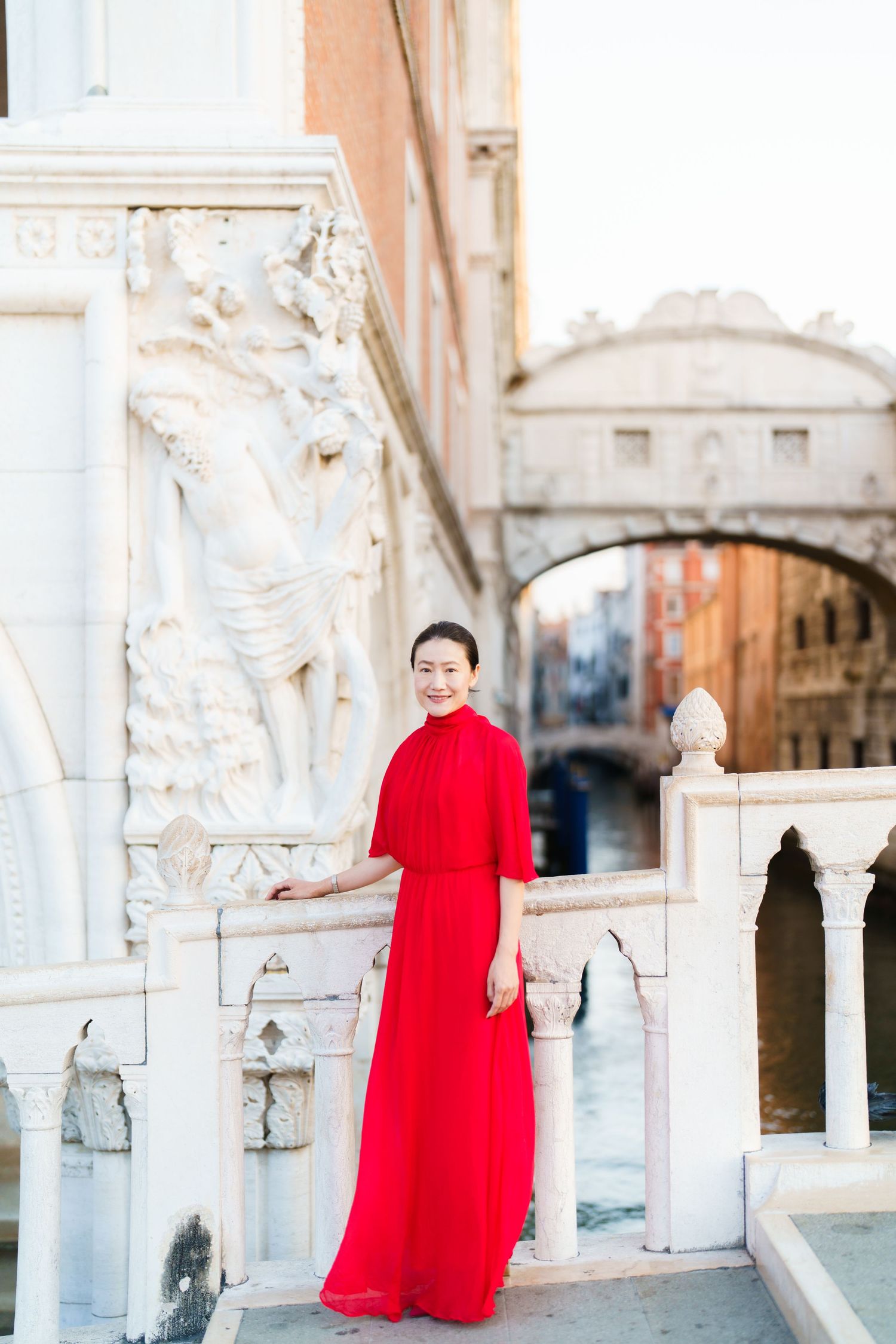 Woman in red gown poses by ornate white marble balustrade with Bridge of Sighs in Venice, Italy.