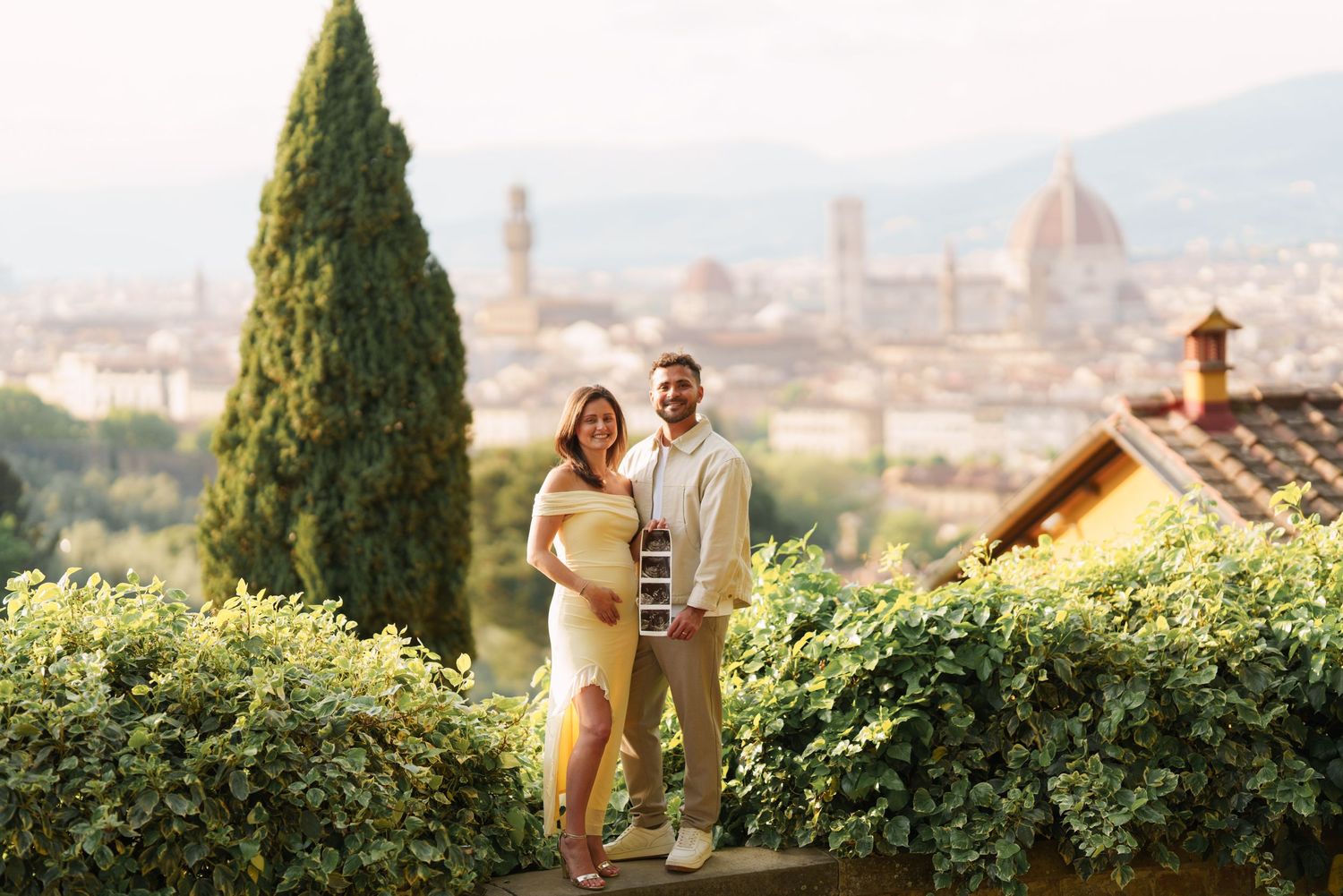 Pregnant couple posing in front of Florence skyline with Duomo cathedral and cypress trees in background.