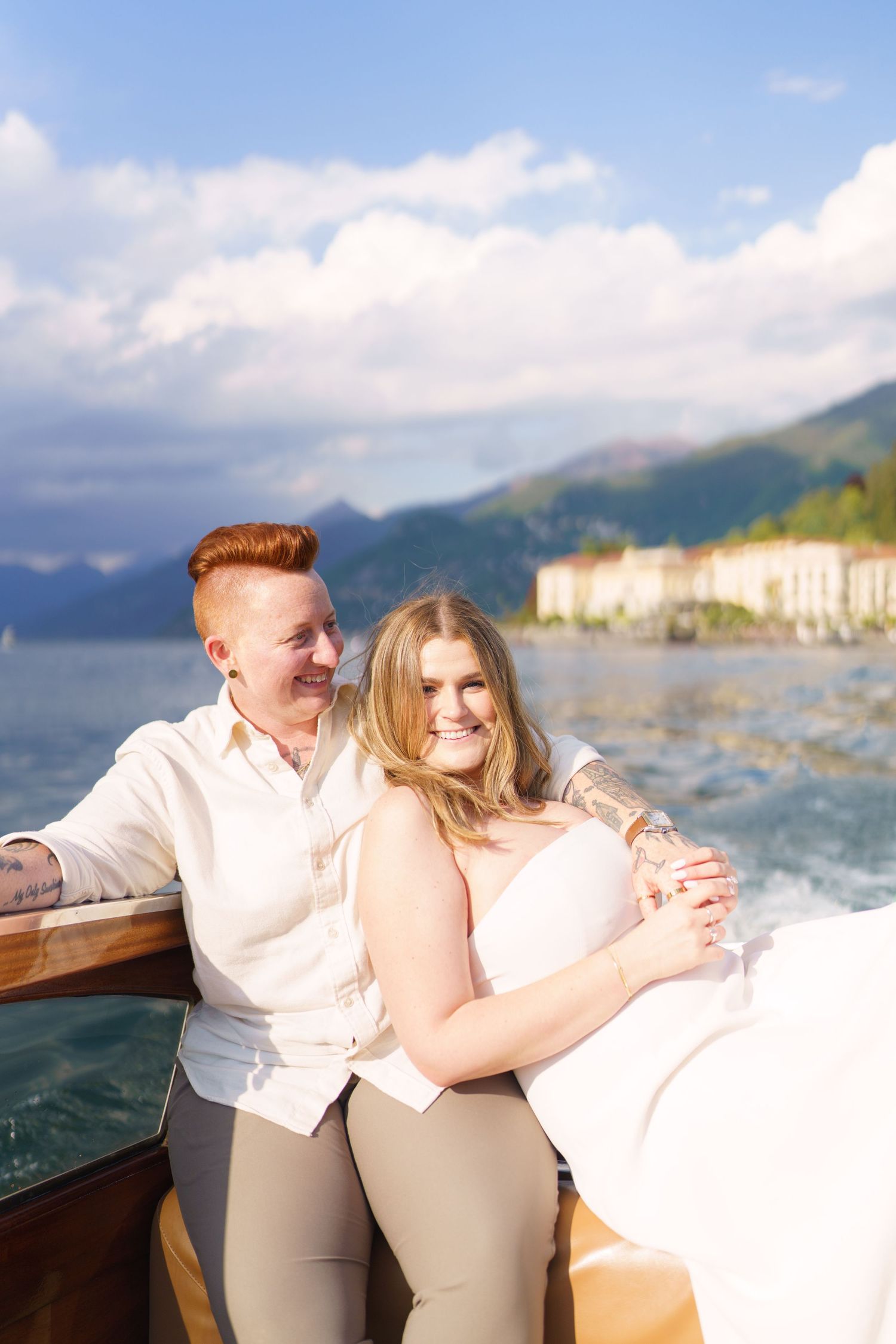 Two women celebrate on a boat with champagne, scenic lake and mountains in the background on a sunny day.