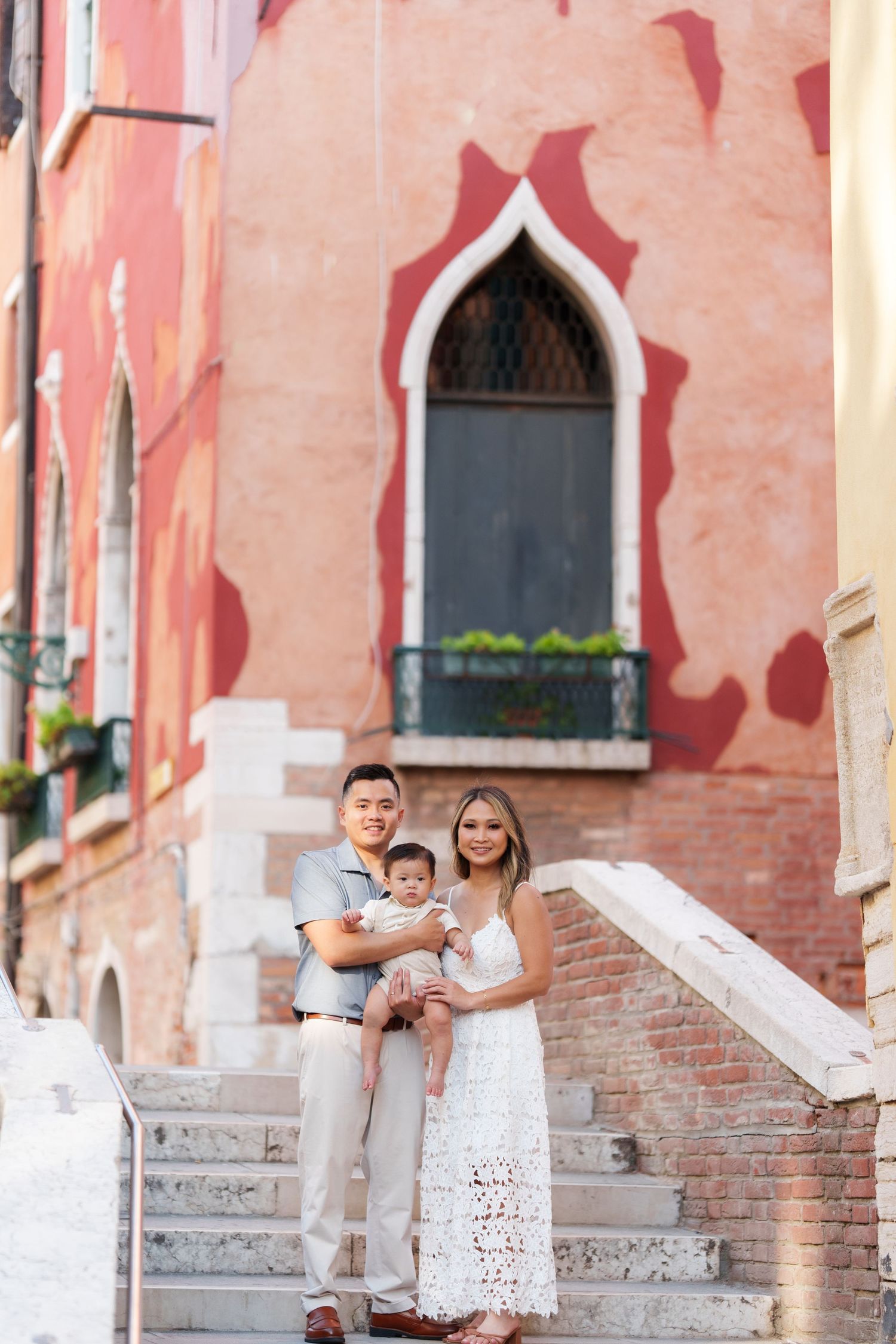 Young family with baby posing on steps before a red Venetian building in Venice, Italy.