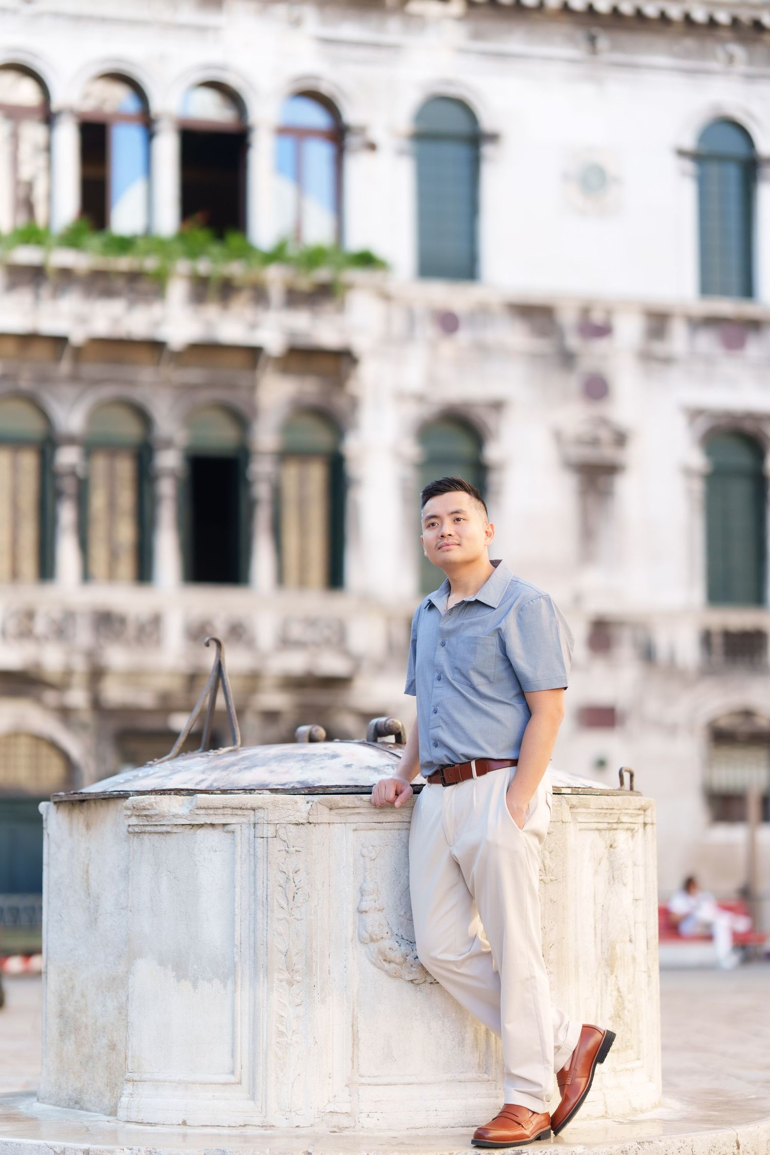 Young Asian man in blue shirt and beige pants leaning against a stone well in a historic European plaza.