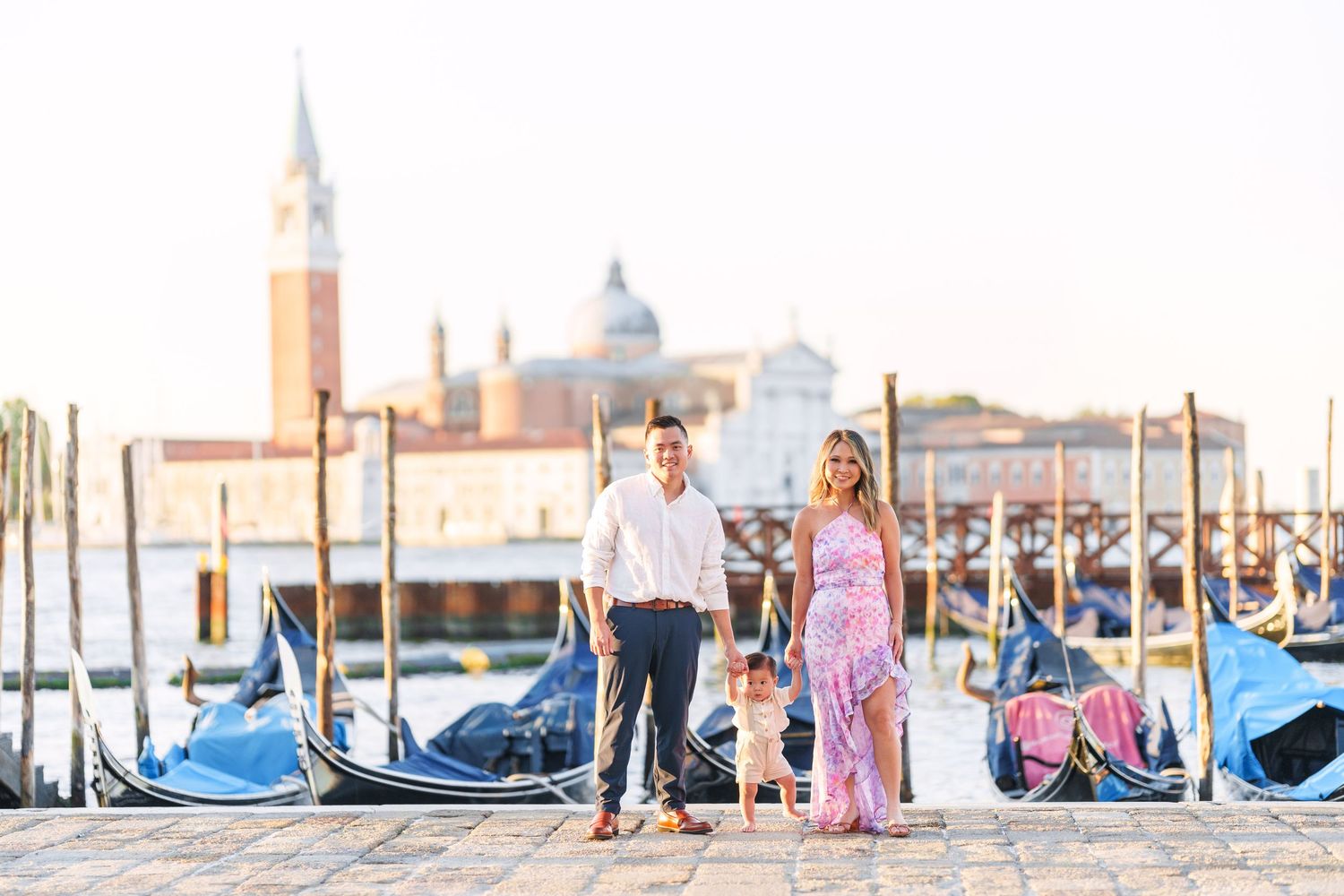 Couple holding hands on Venice waterfront with gondolas and San Giorgio Maggiore church at golden hour.