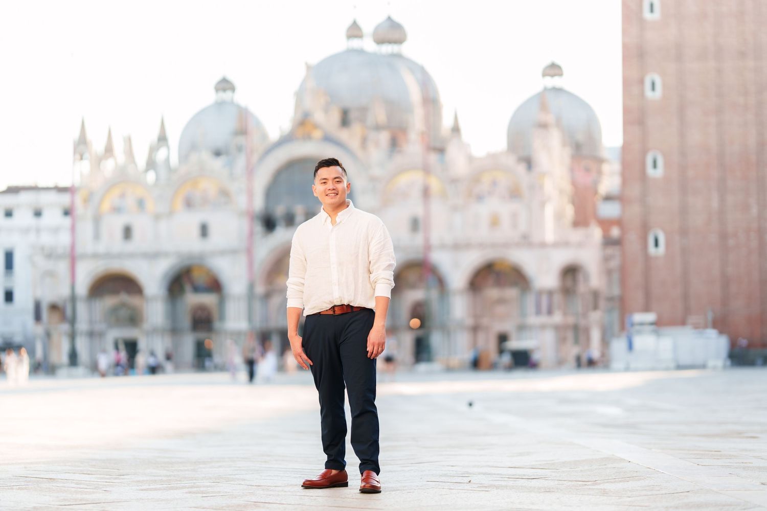 Man in white shirt and navy pants stands in St. Mark's Square, Venice, with the iconic Basilica in the background.
