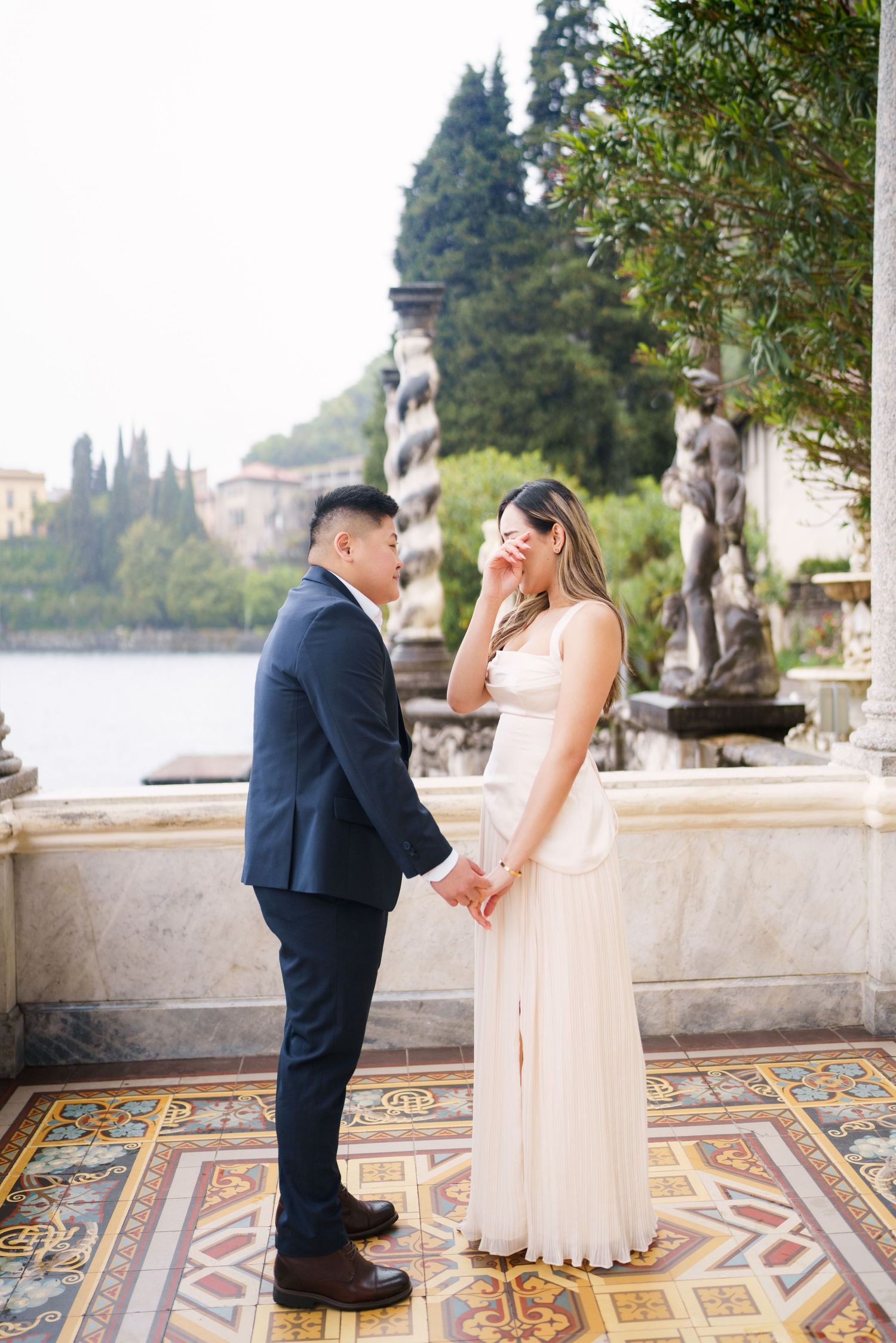 Elegant couple photography at Villa Monastero in Varenna by Lake Como propohite gown wiping tears, man in navy suit, Italian villa backdrop.