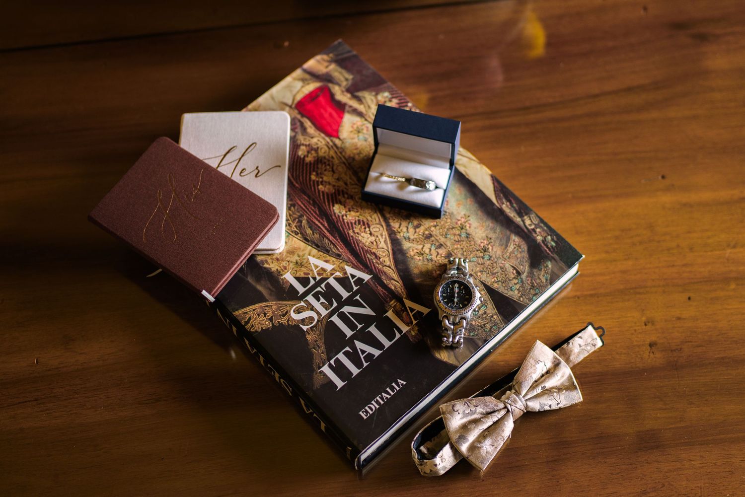 Elegant groom accessories flat lay featuring cufflinks, watch, bow tie, and books on a wooden surface.