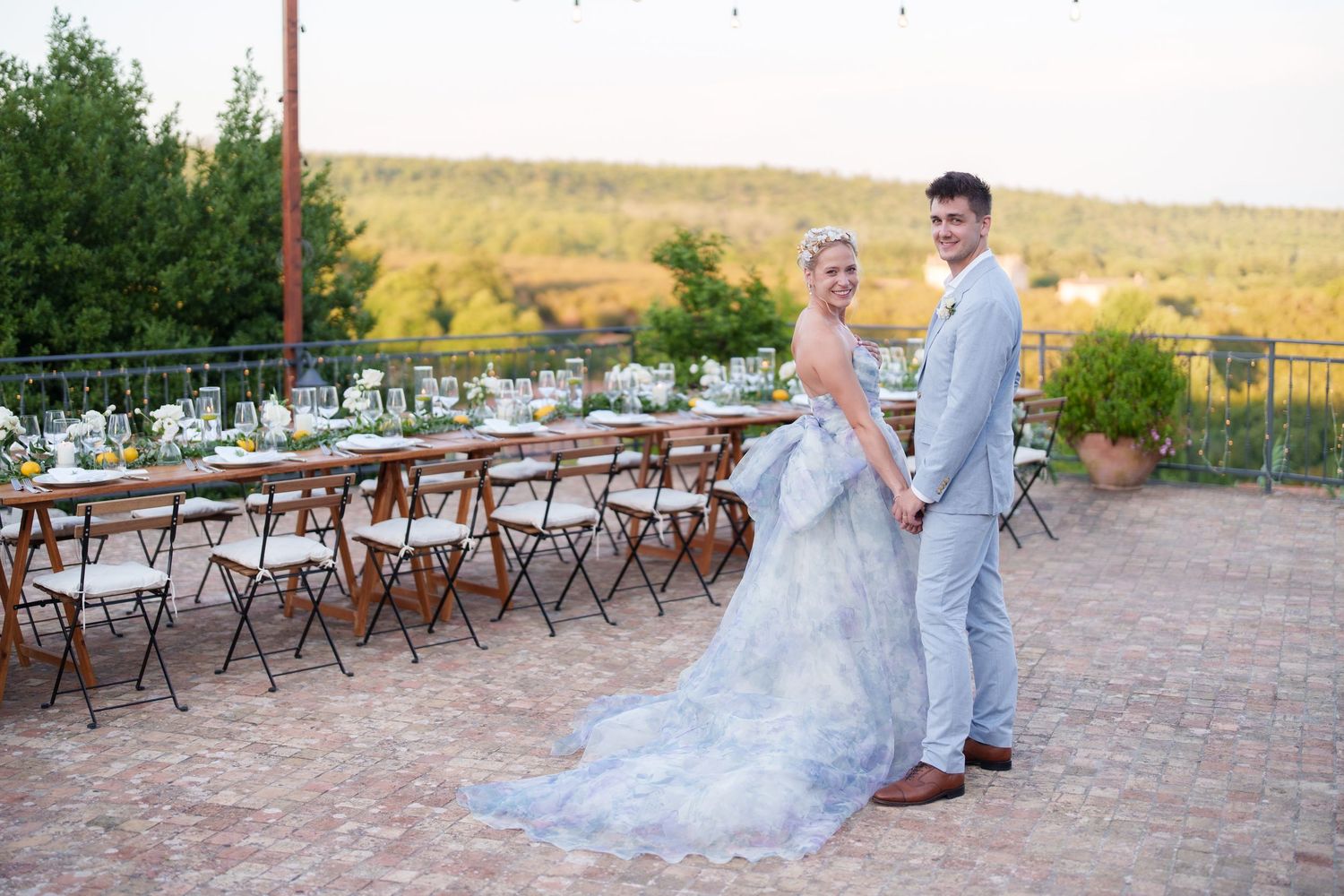 Bride and groom holding hands on a brick patio with elegant outdoor reception tables set against a scenic landscape.