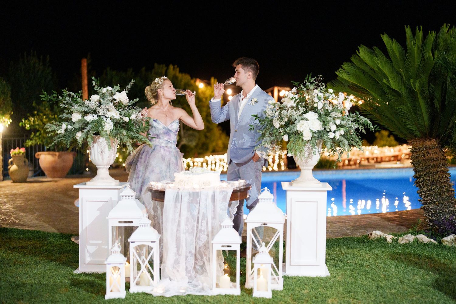 Couple toasting champagne at elegant outdoor wedding reception with white floral arrangements and lanterns by pool at night.