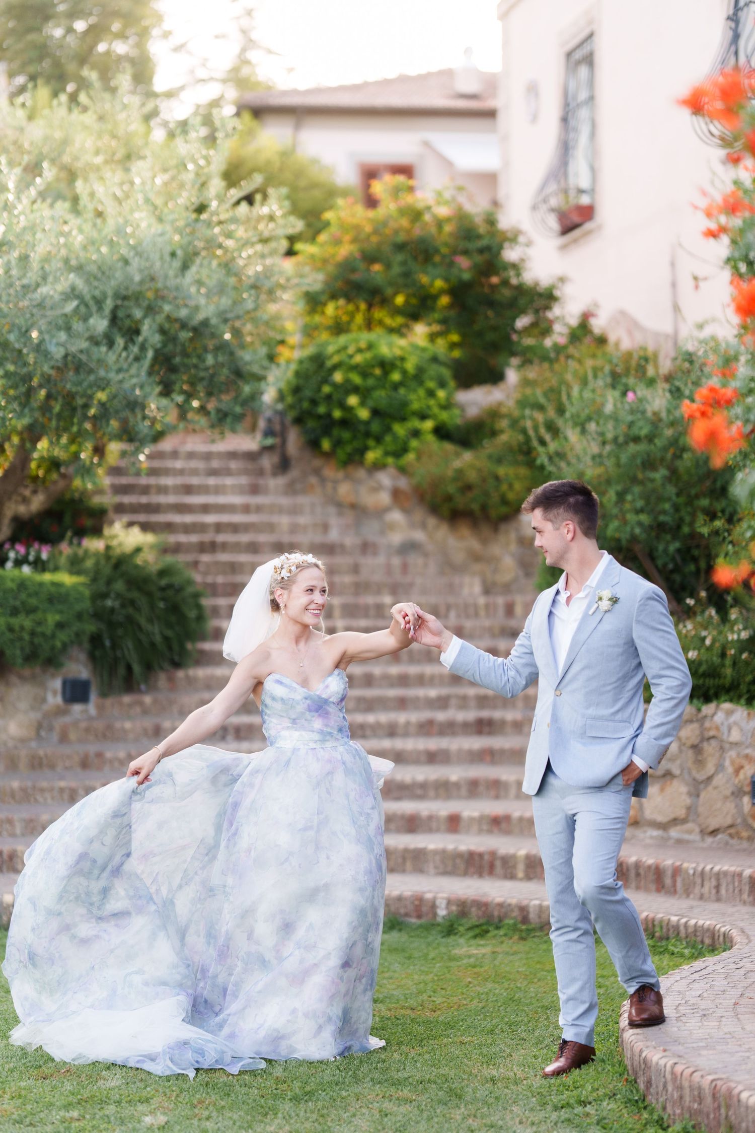 Bride in blue watercolor gown holds hands with groom in light gray suit dancing in lush garden with stone steps.