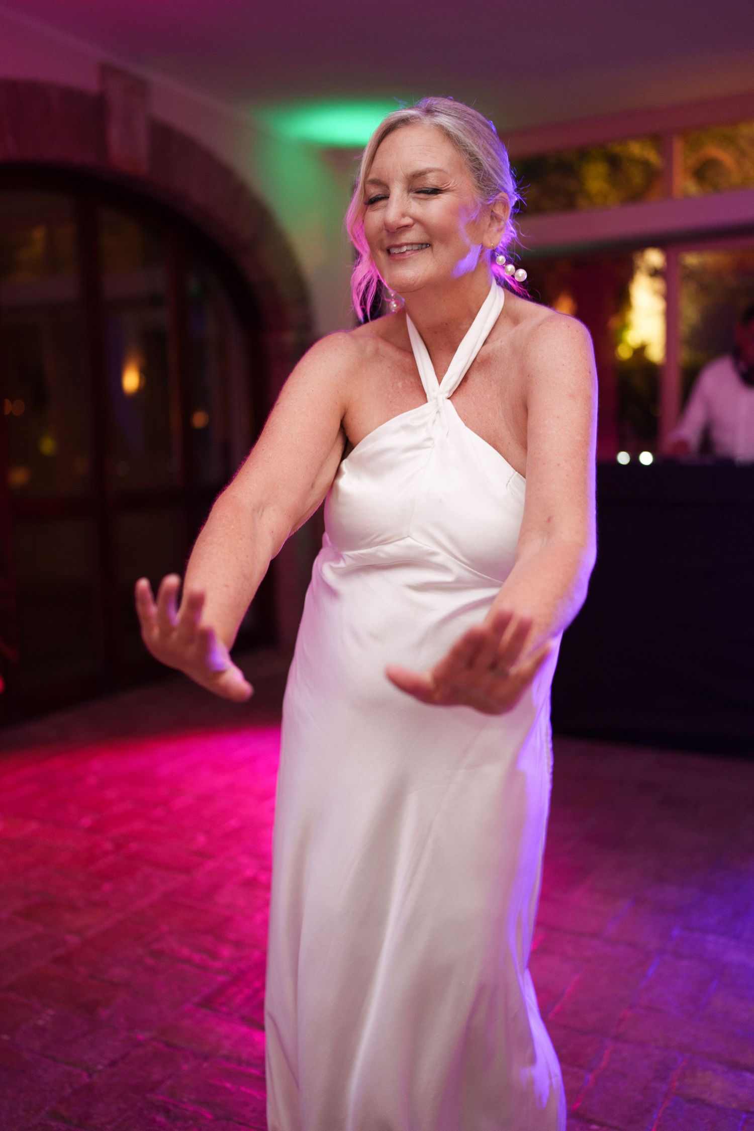 Bride in white halter gown dancing joyfully at wedding reception with colorful pink and green lights.