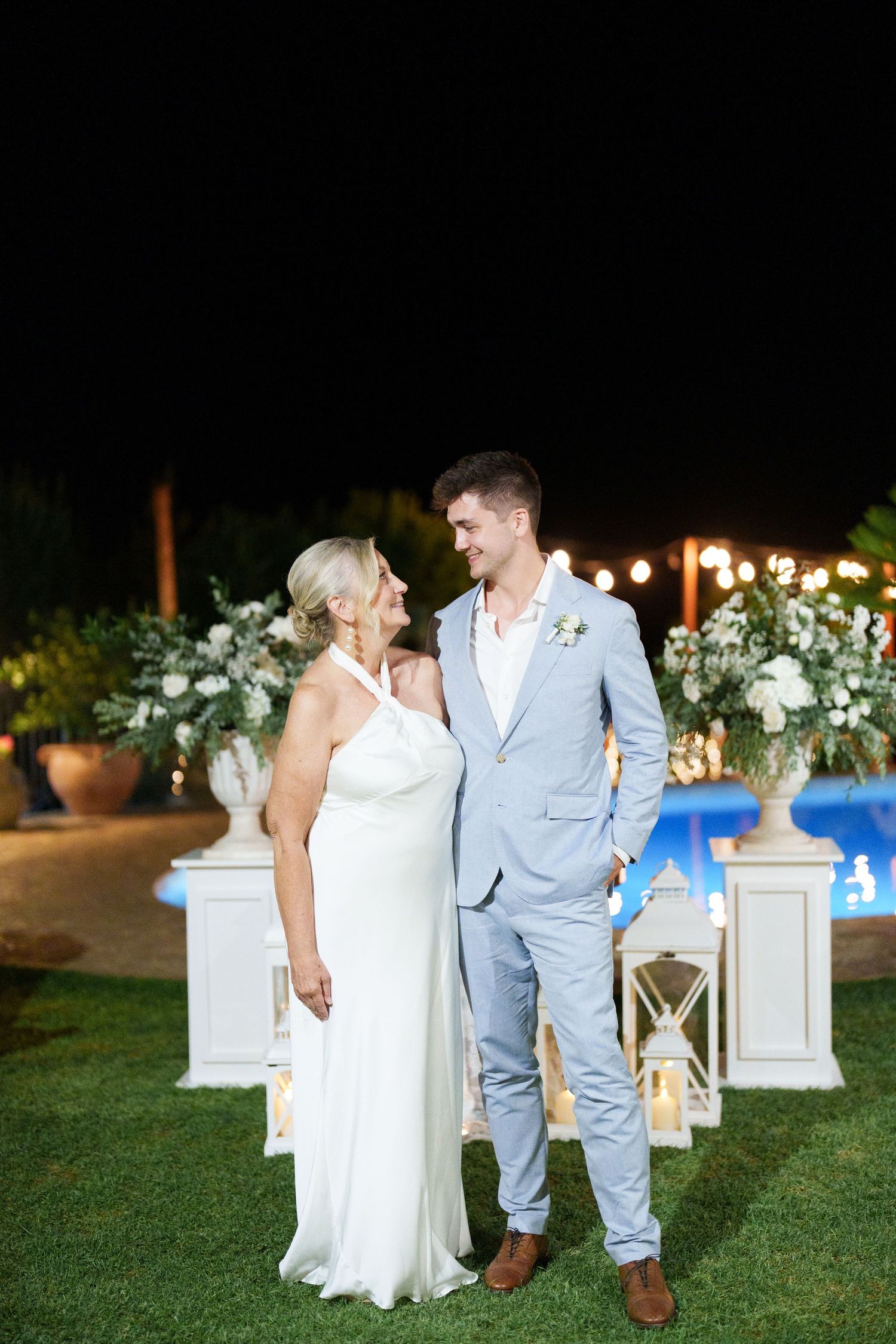 Bride in white halter gown and groom in light blue suit pose together at night wedding reception near pool.