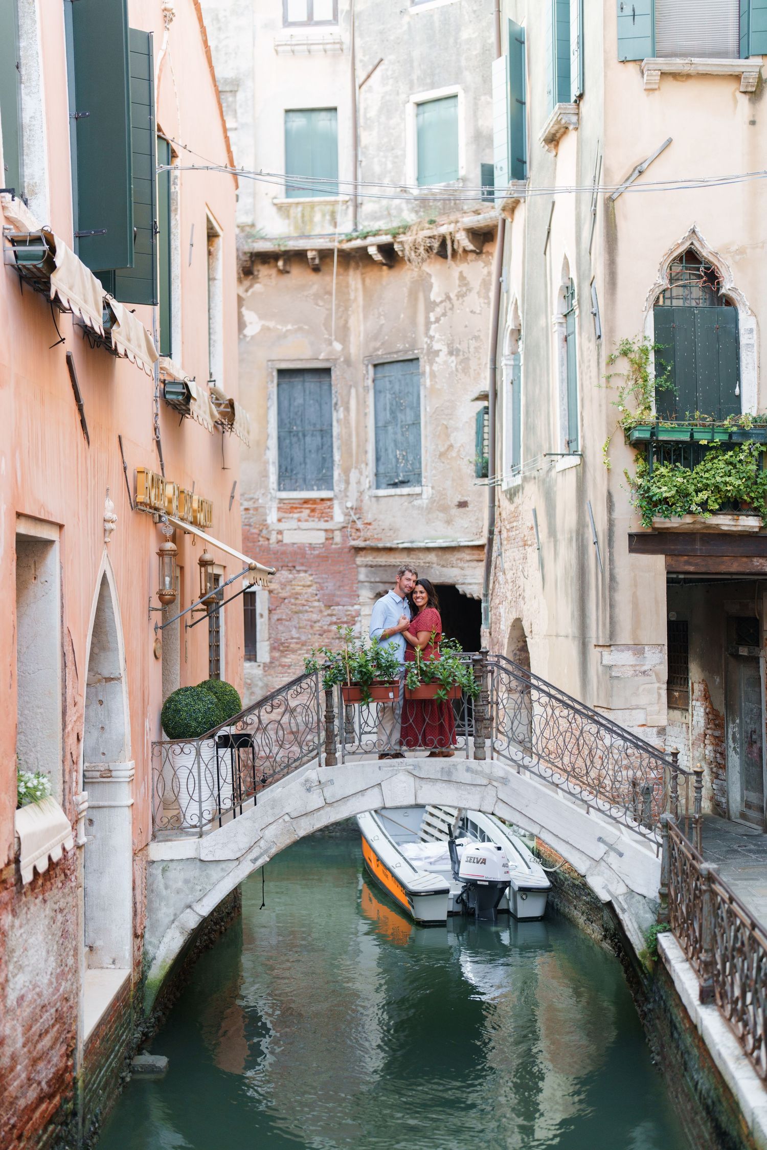 Couple embracing on a stone bridge over a Venice canal, surrounded by colorful historic buildings and moored boats.