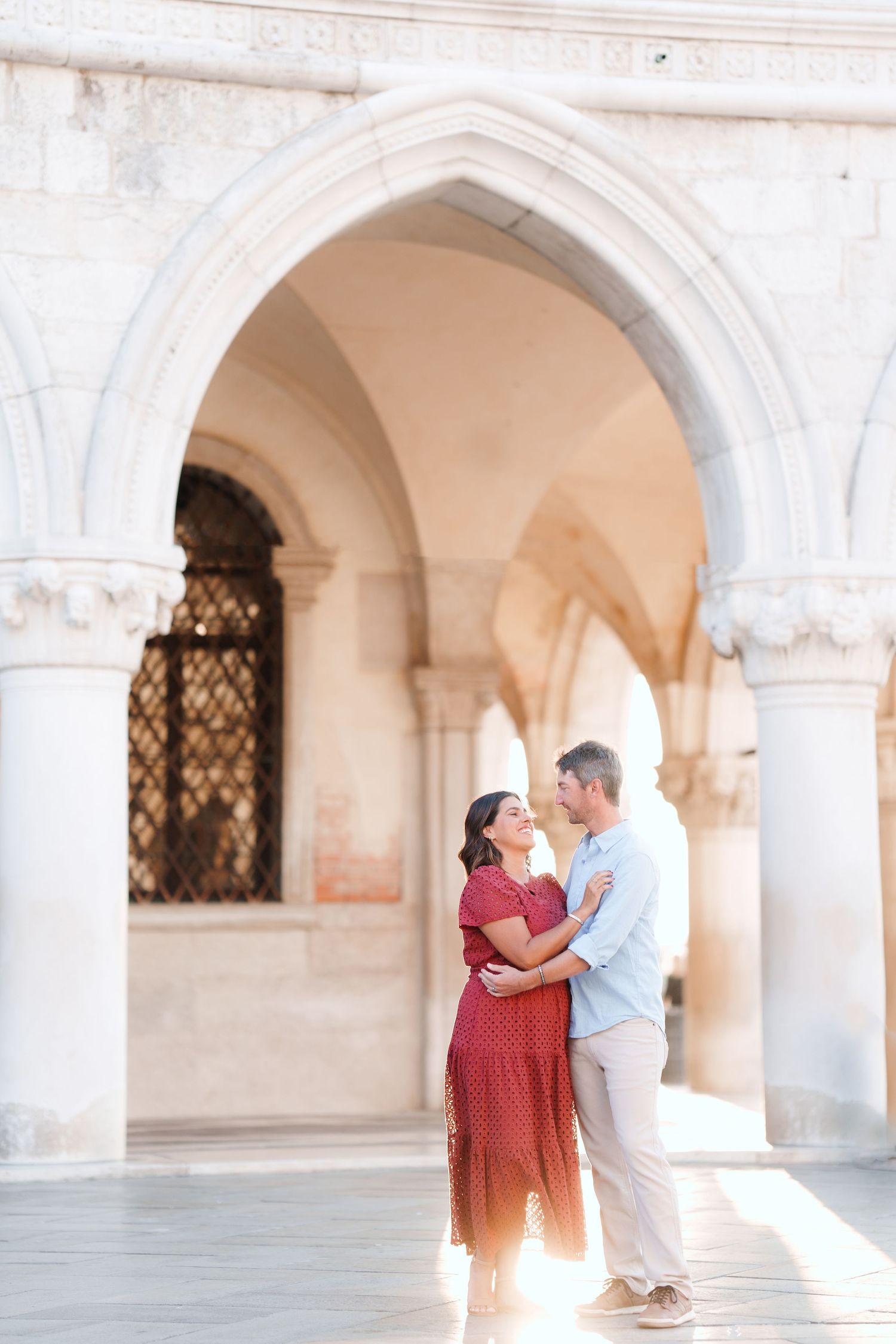Couple embracing under ornate Gothic arches in Venice, woman in red dress, man in blue shirt, warm sunlight.