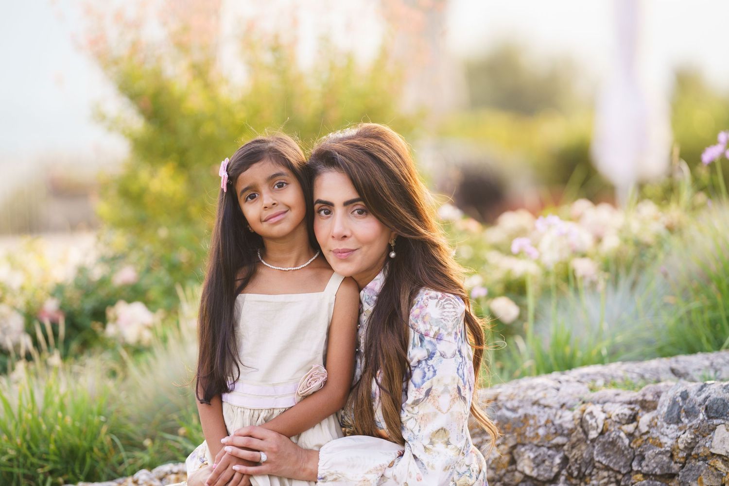 A woman and young girl pose together outdoors in a sunlit garden, wearing white outfits for a family portrait.