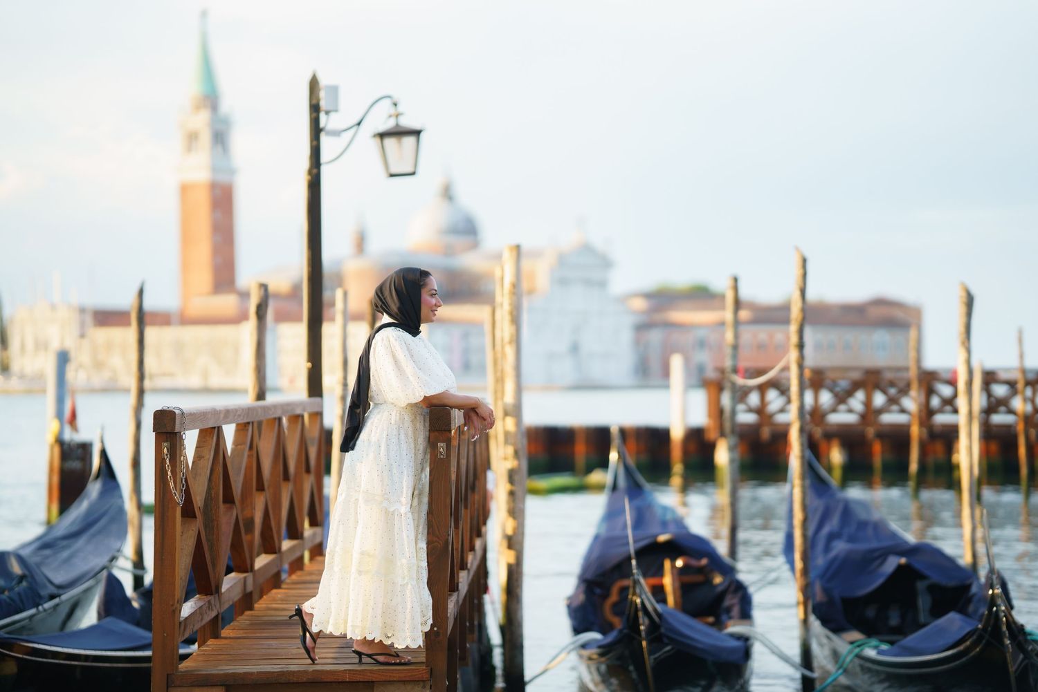 Woman in white dress standing on a wooden dock in Venice with gondolas and San Giorgio Maggiore in background.