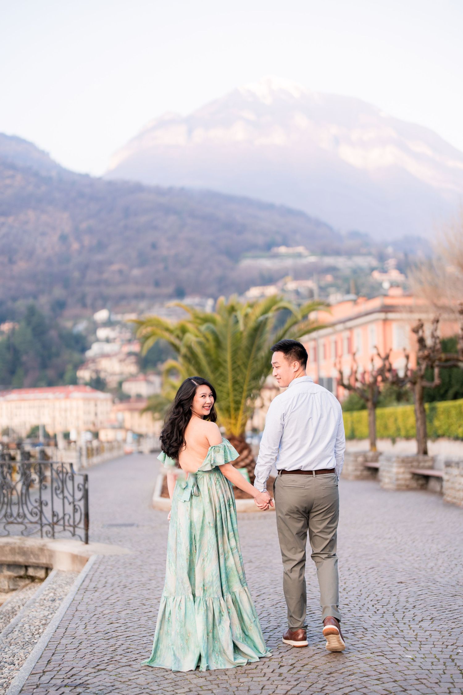 Couple holding hands on cobblestone path with mountain backdrop, woman in green floral dress, man in casual attire.
