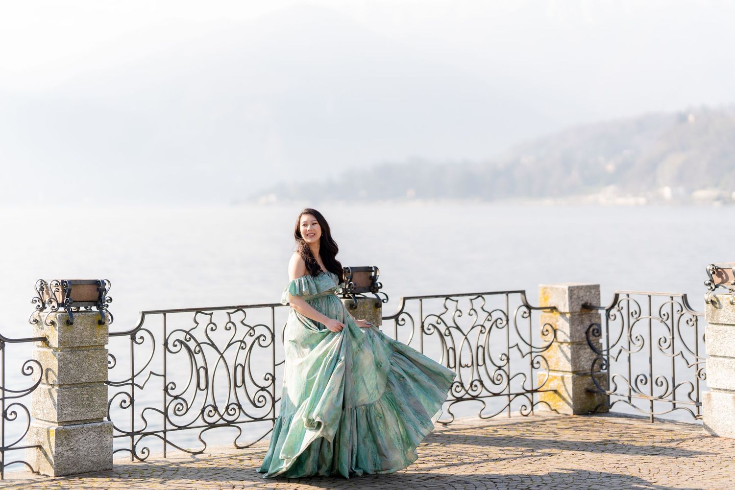 Woman in flowing teal gown poses on ornate iron terrace overlooking misty lake and mountains.