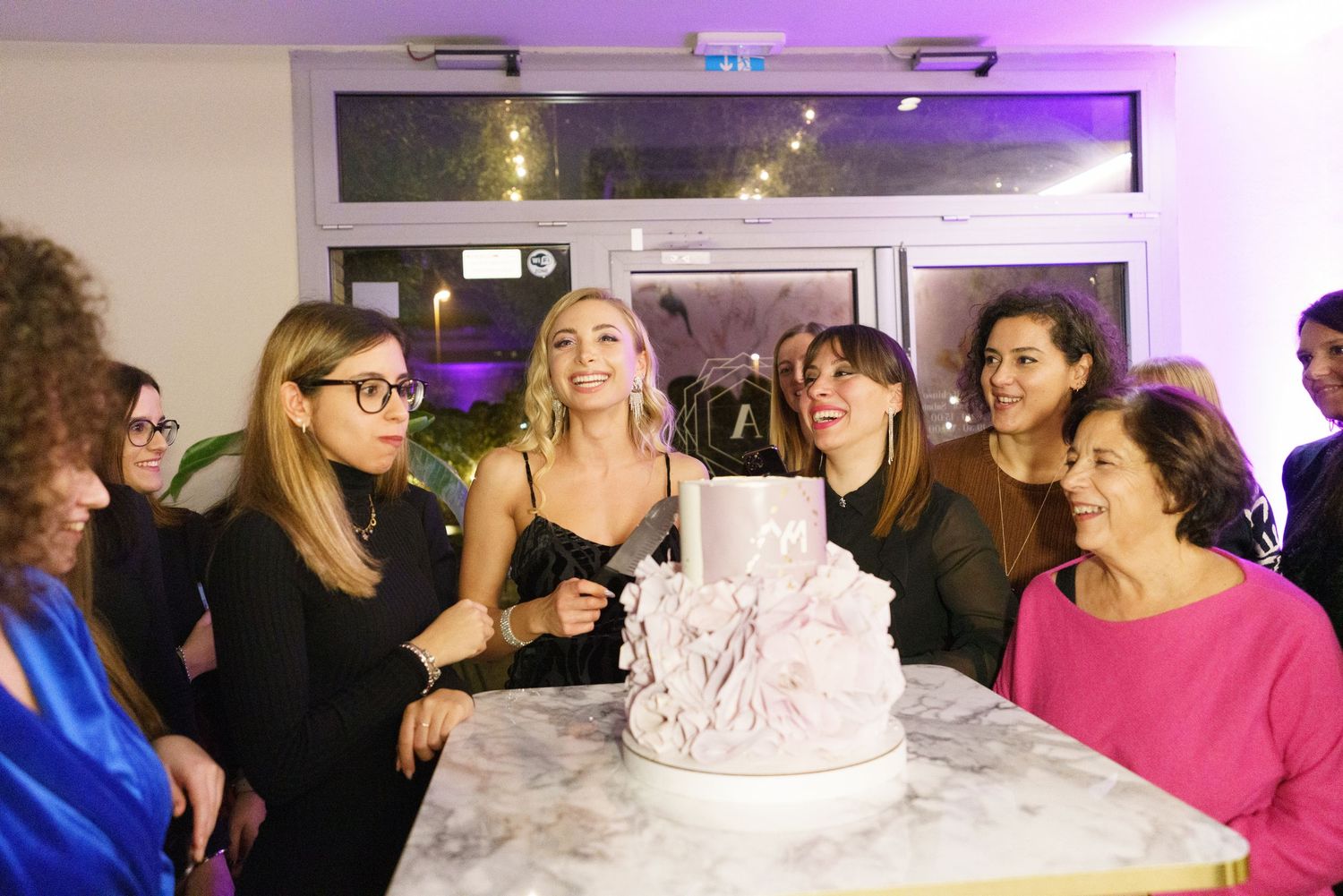 Group of women gathered around a white tiered celebration cake at an indoor party event.
