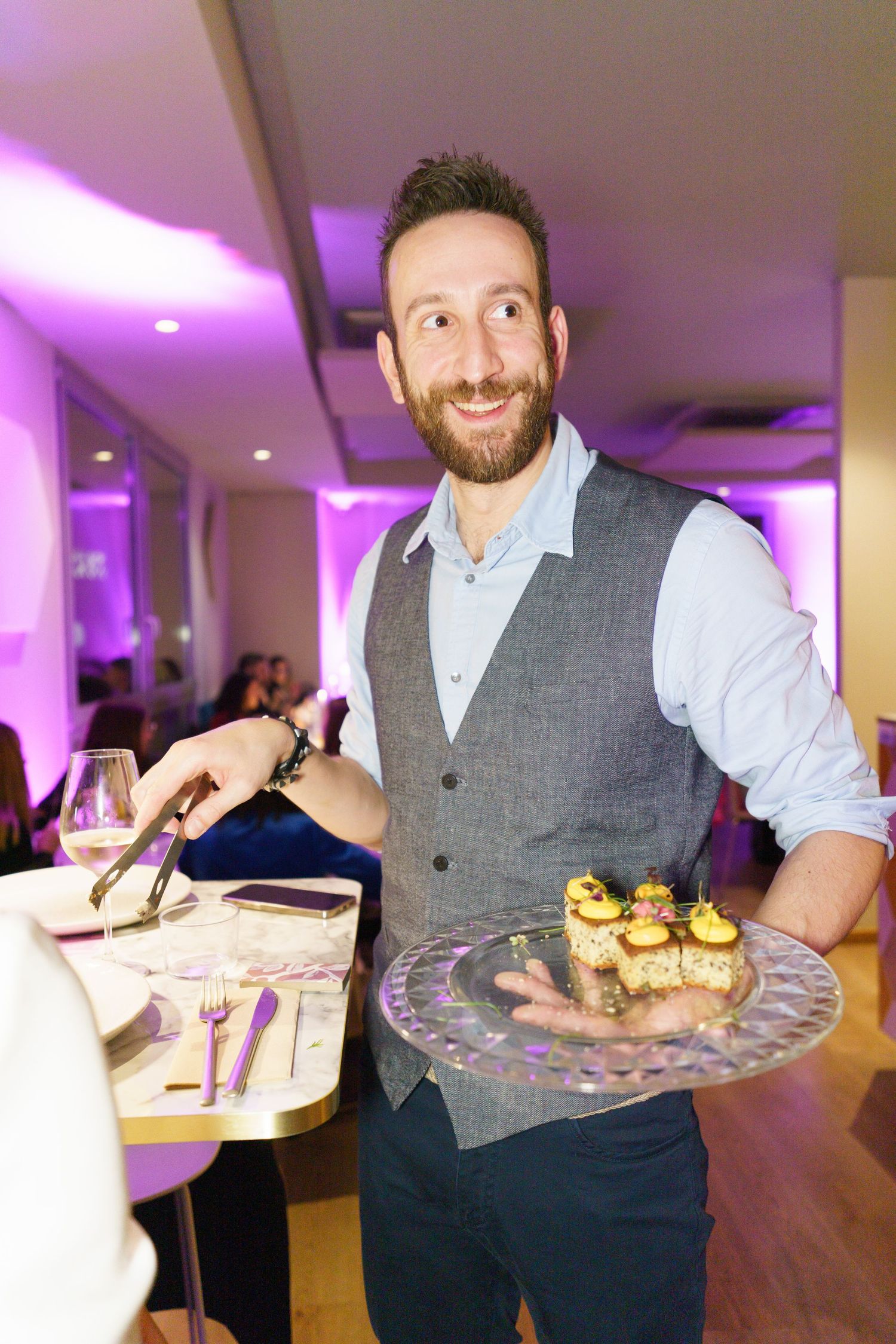 Smiling bearded waiter in gray vest holding a tray of appetizers in a purple-lit restaurant.