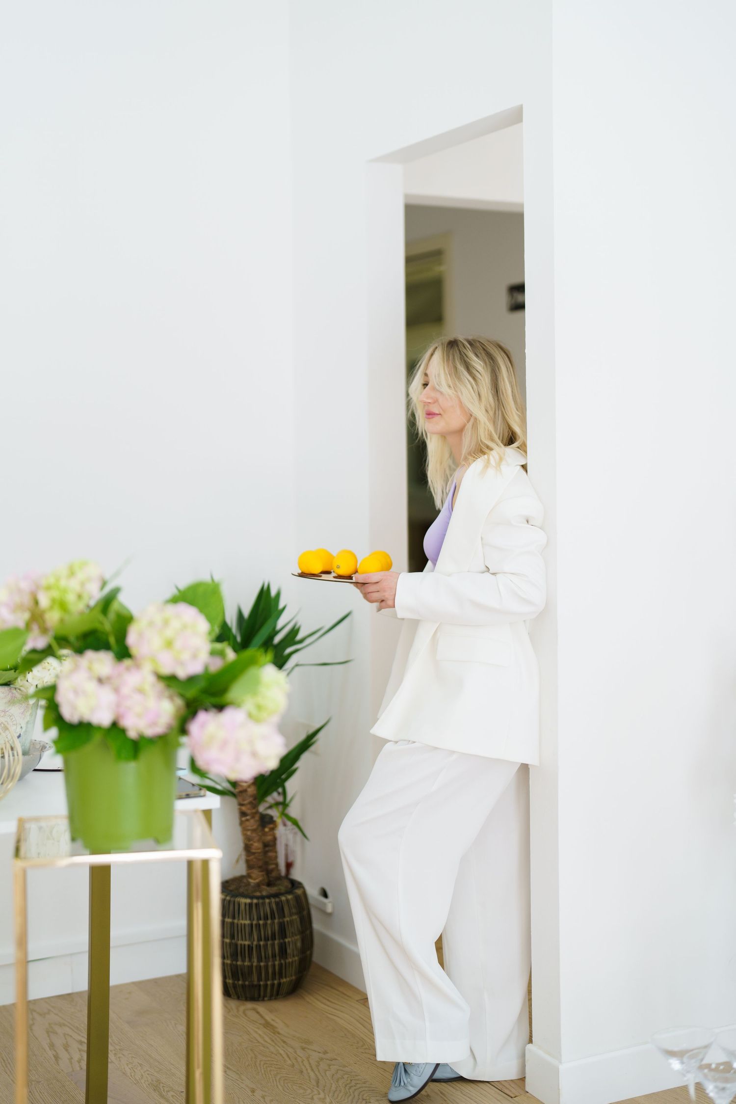 Blonde woman in white suit holding oranges, standing in doorway near floral arrangement in bright modern interior.