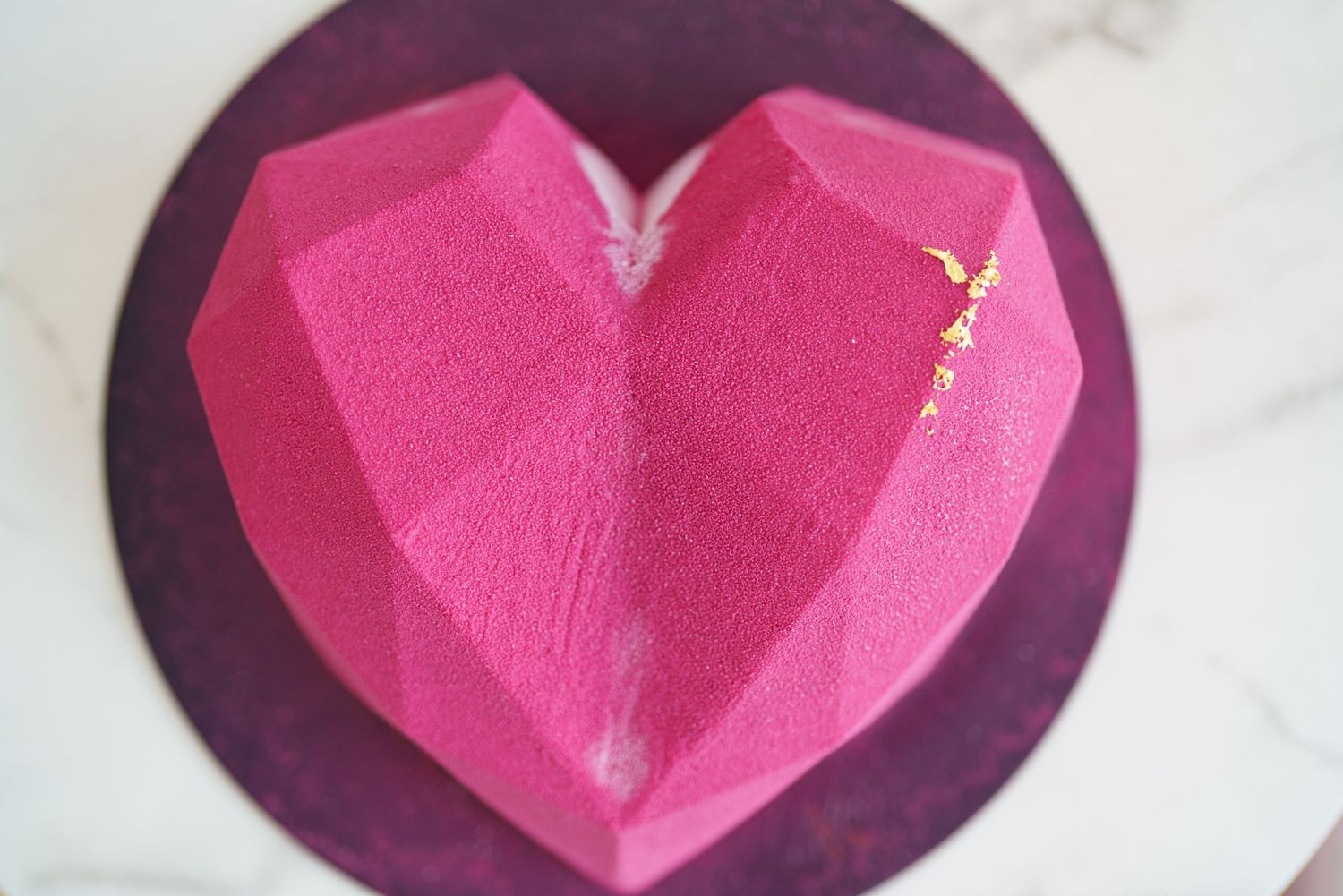 Geometric pink heart-shaped cake with gold leaf accents on a purple board, perfect for Valentine's Day celebrations.