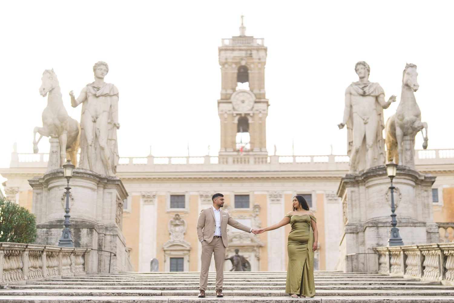 Couple holding hands on Capitoline Hill steps with classical statues and Palazzo Senatorio in Rome, Italy.