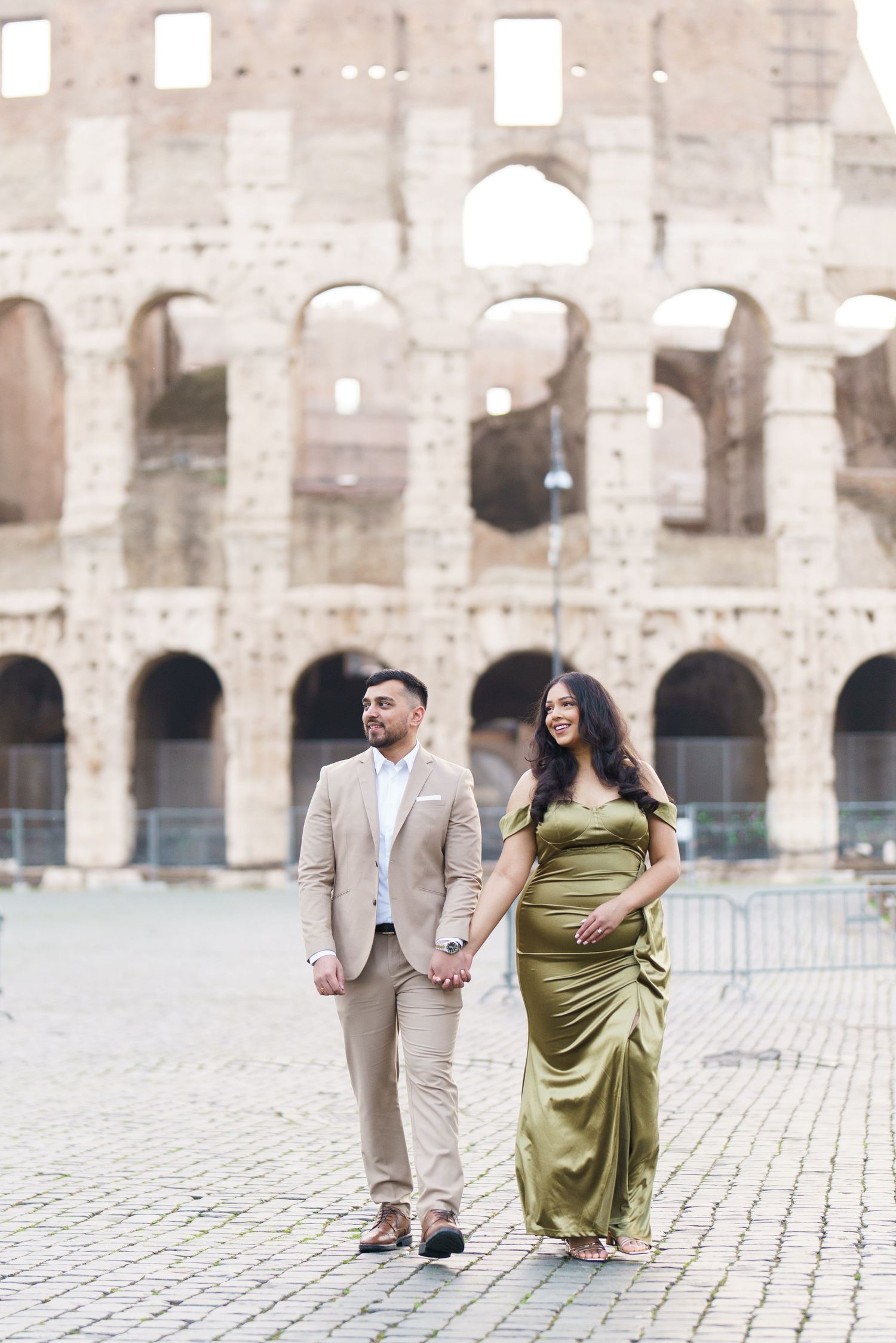 Couple holding hands in front of the Colosseum in Rome, woman in olive gown, man in beige suit.