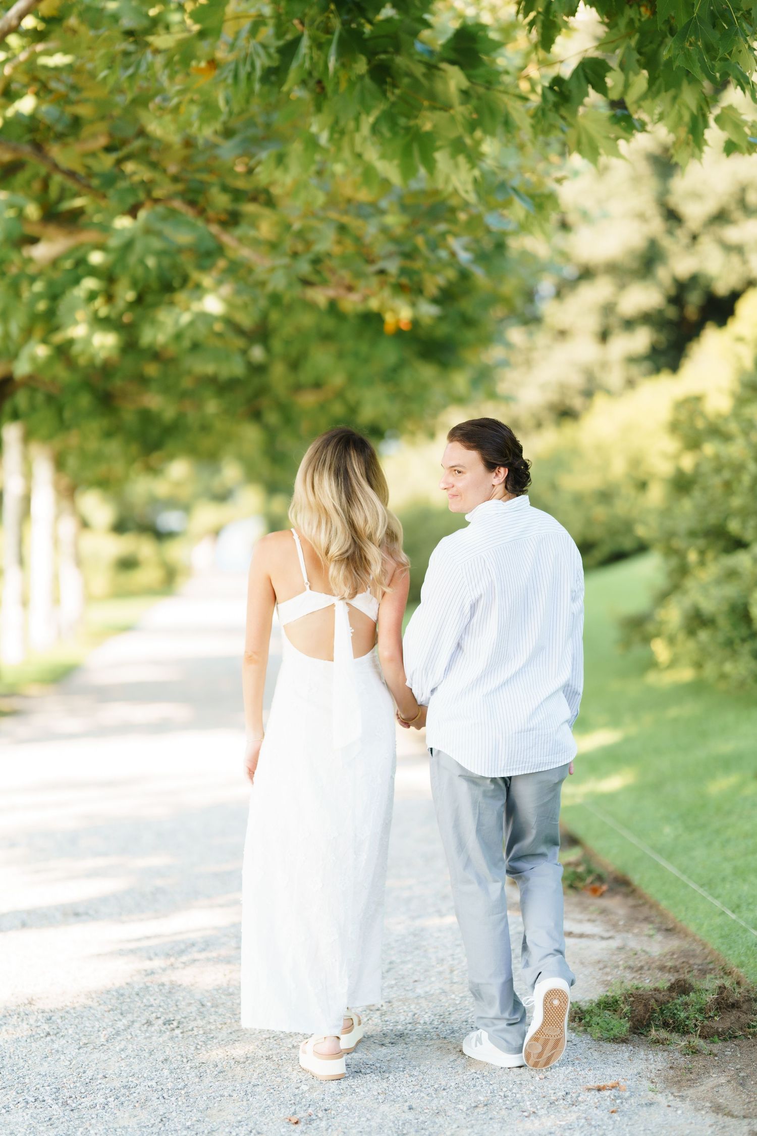A couple in white and blue outfits walk together on a tree-lined path during a romantic outdoor photo session.