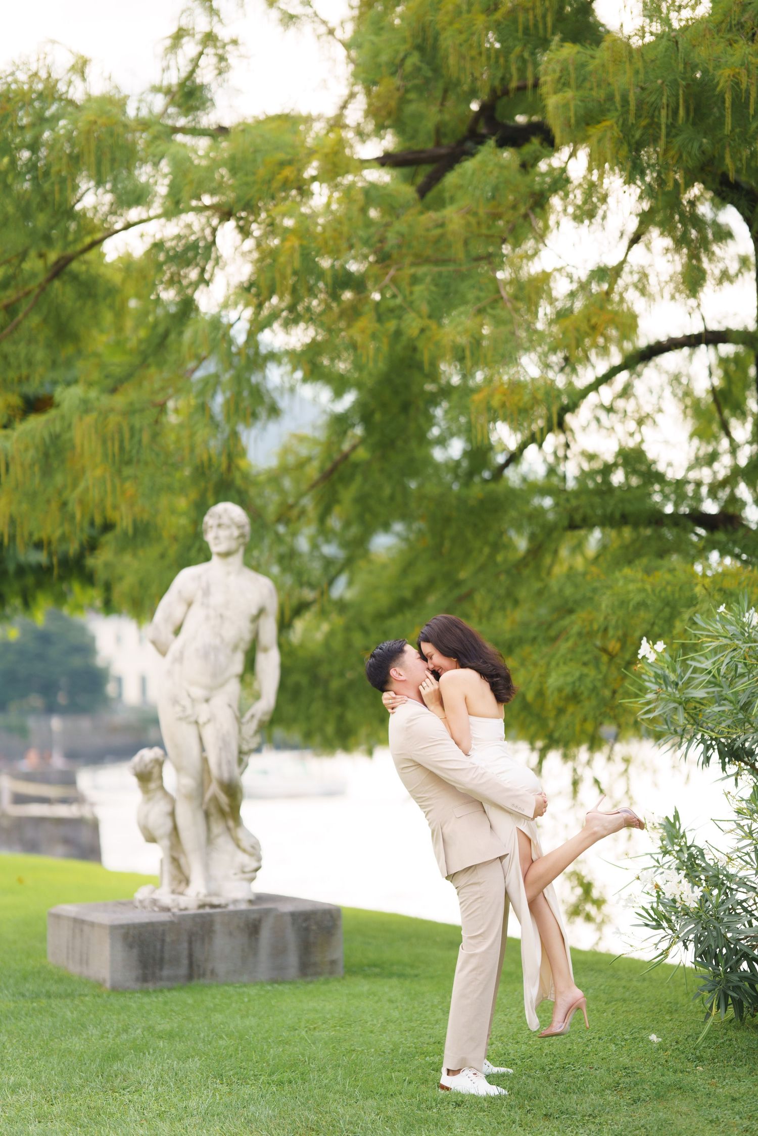 A couple embraces on a grassy lawn with stone statues in the background on a sunny summer day.